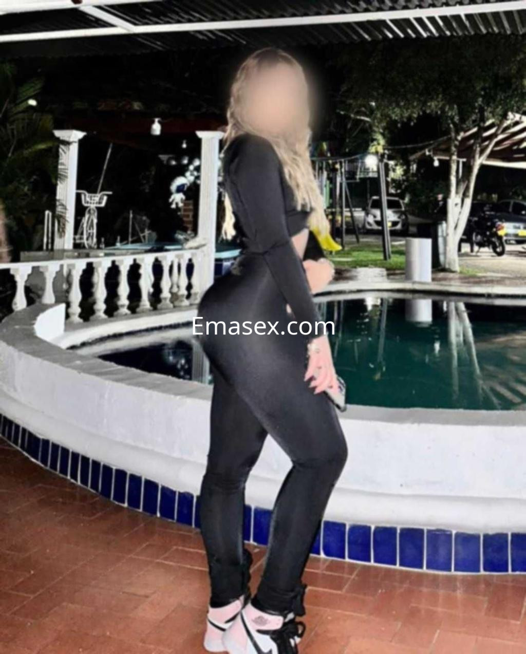Escort en Valladolid, vistiendo un conjunto negro ajustado, posa junto a una piscina en un ambiente al aire libre.