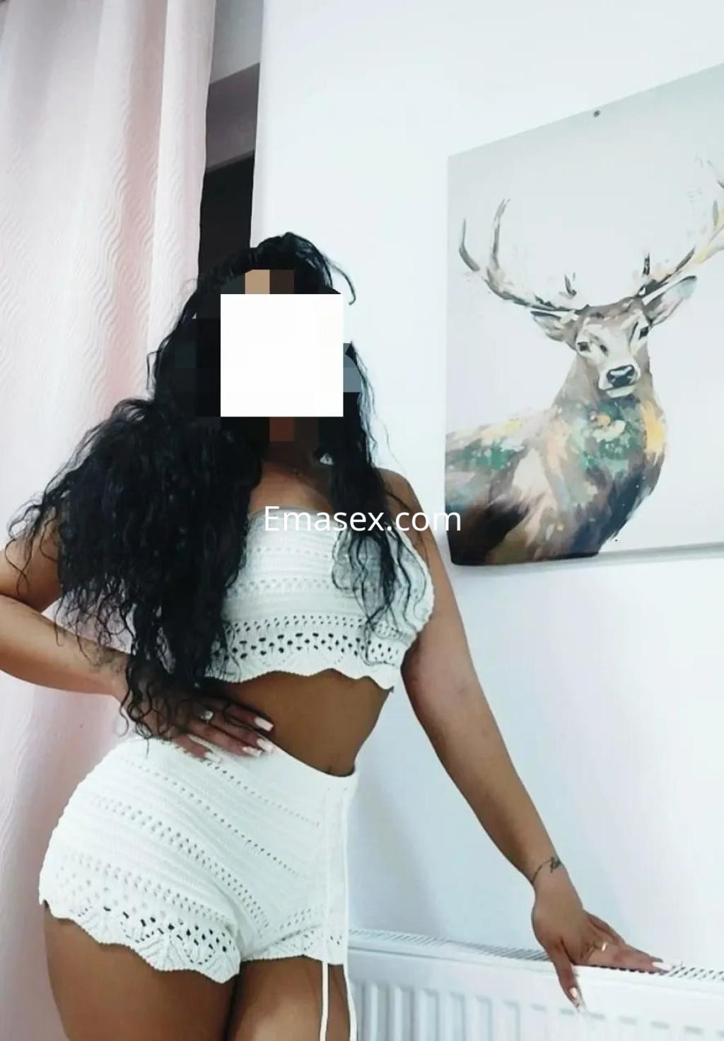 La puta viste un conjunto blanco de crochet, posando con una mano en la cadera en un ambiente decorado en Valladolid.