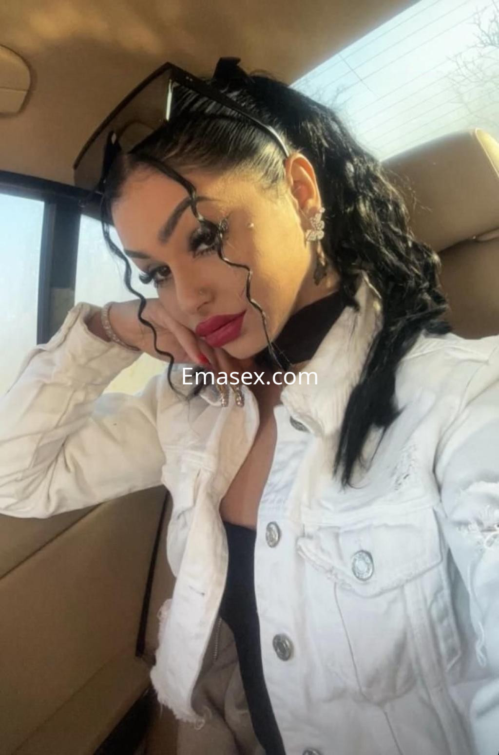 Escort en Bilbao con cabello oscuro y rizado, usando chaqueta blanca y gafas de sol, sonríe en el asiento de un coche.