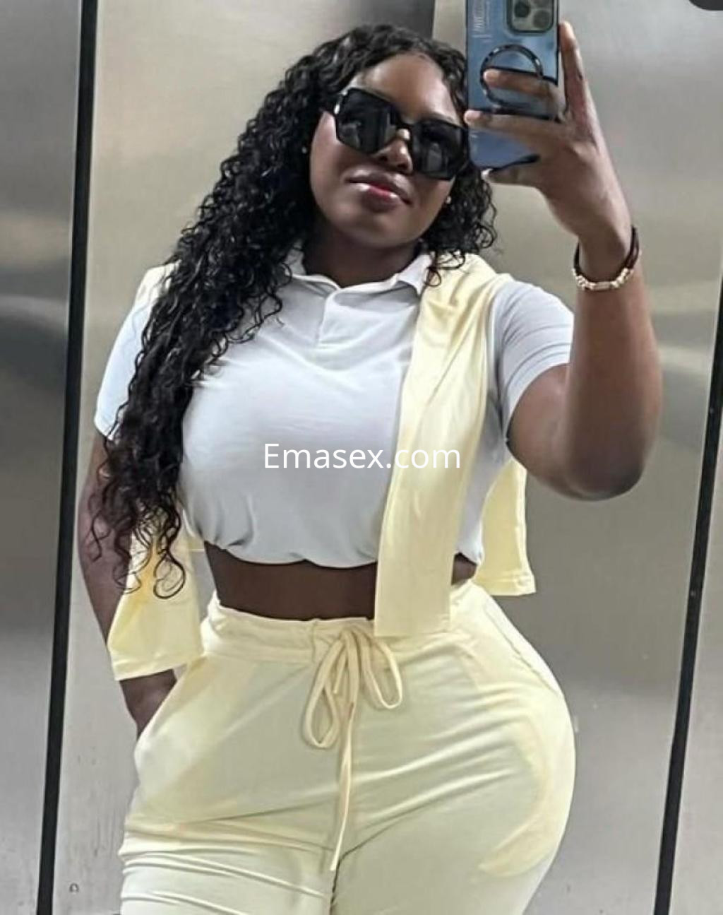 Escort en Zaragoza con cabello rizado y oscuro, viste un conjunto amarillo claro y gafas de sol en un espejo.
