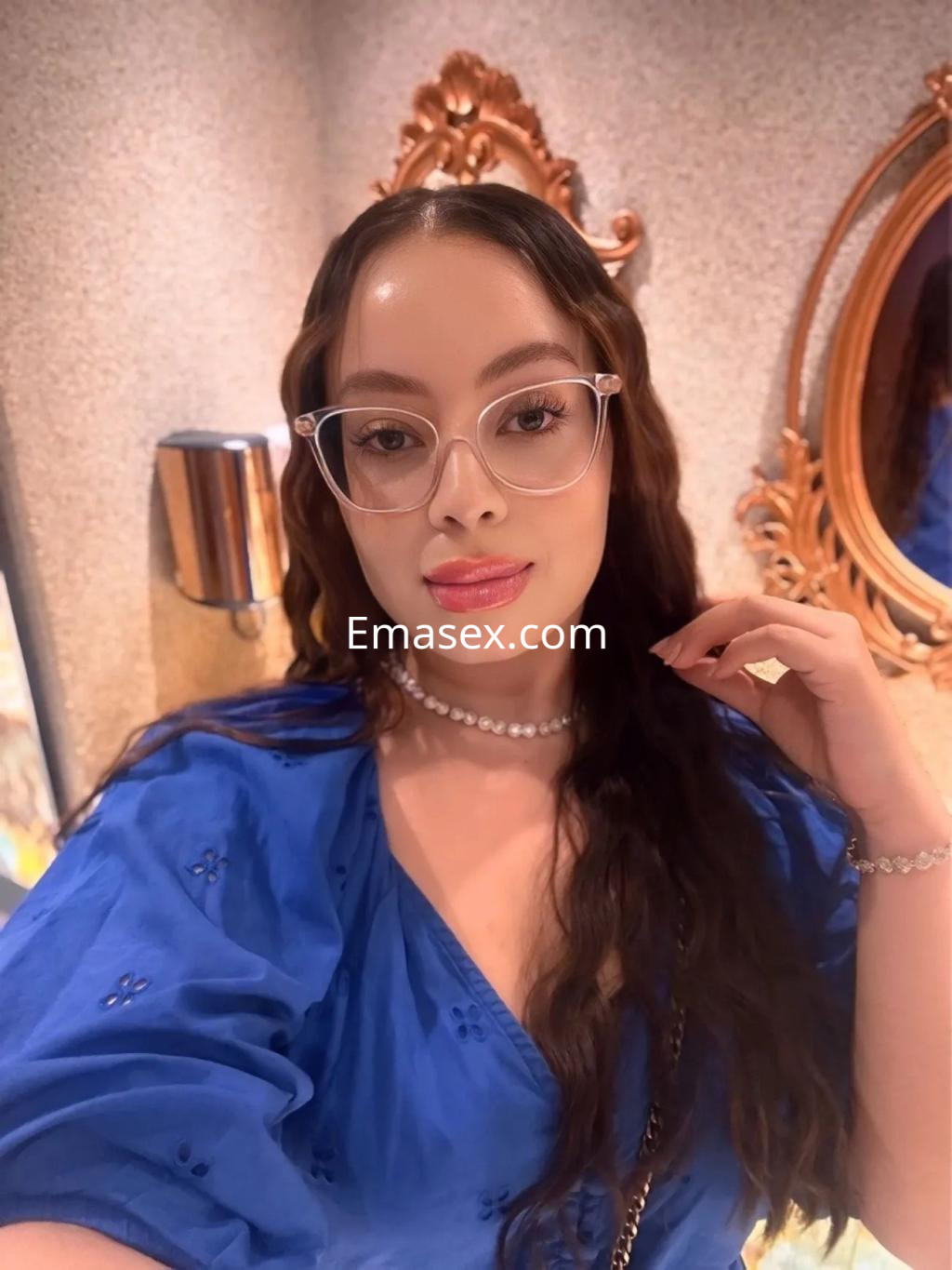 Escort en Zaragoza, con blusa azul, cabello largo y ondulado, luciendo gafas y un maquillaje sutil.