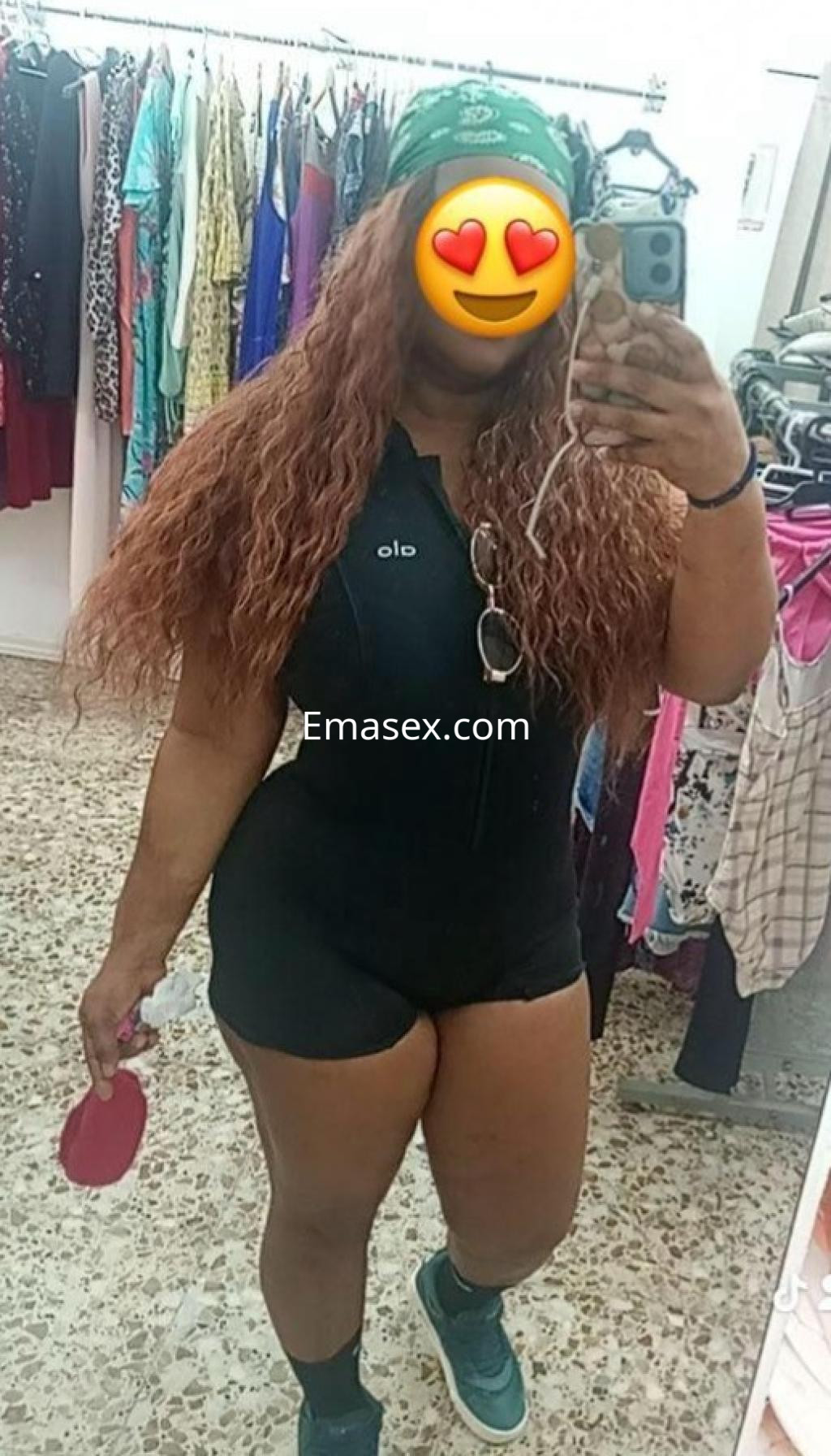 Escort en Albacete posando con un mono negro ajustado, gorra verde y cabello rizado, en un vestidor.