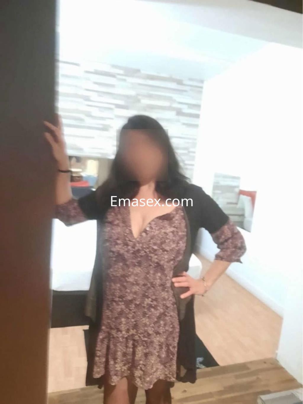 Puta en Barcelona viste un vestido floreado con escote, posando con una mano en la pared en un ambiente acogedor.