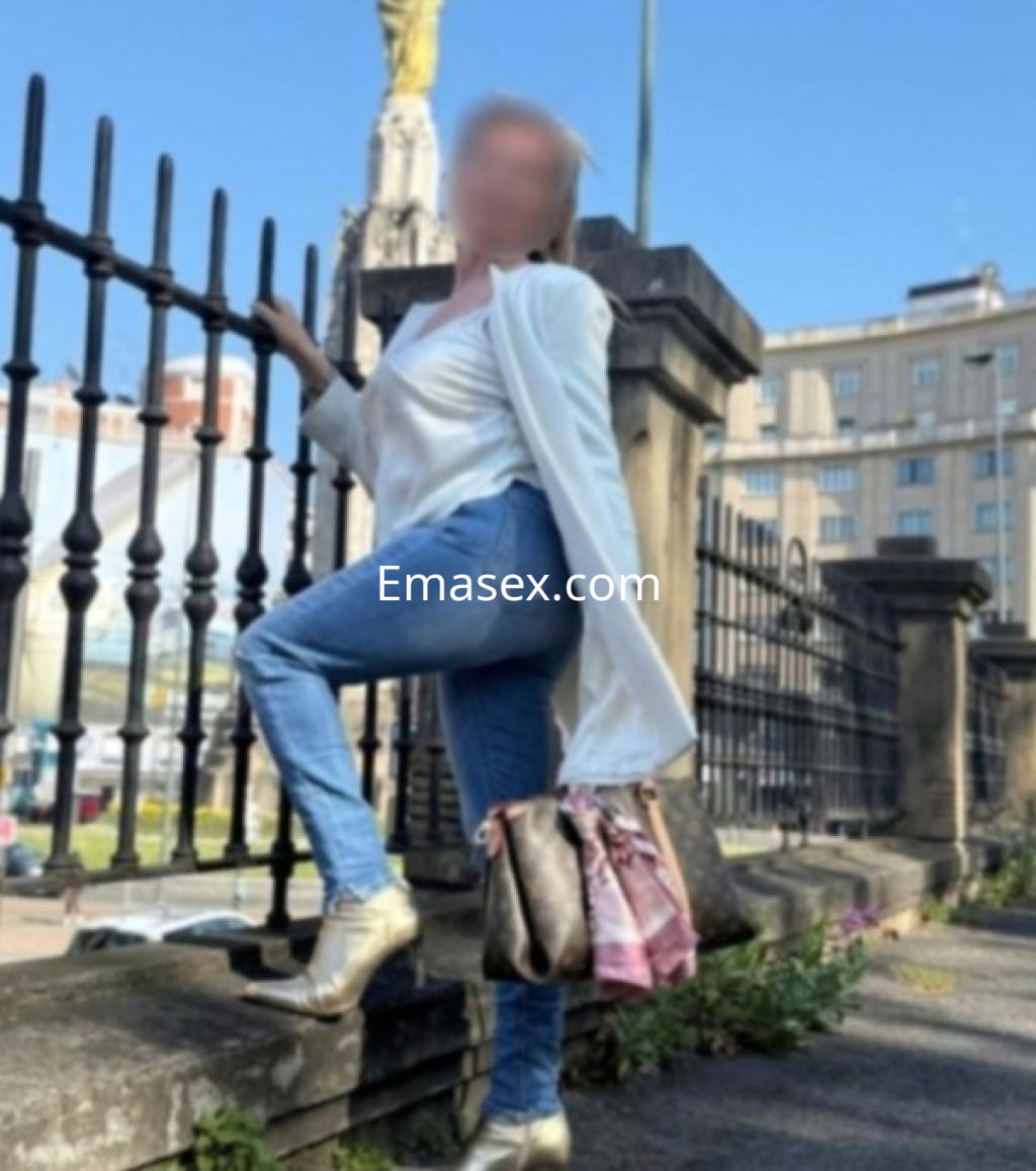 Puta en Coruña, posando en una reja, viste blusa blanca y jeans, con zapatos dorados y bolso en la mano.