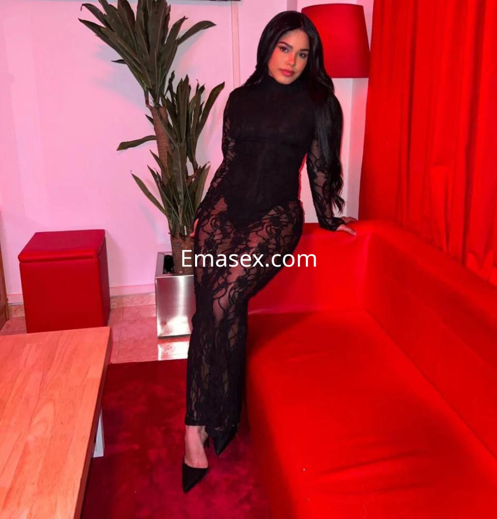 Puta sentada en un sofá rojo, vistiendo un elegante vestido negro de encaje, en un ambiente acogedor en Coruña.