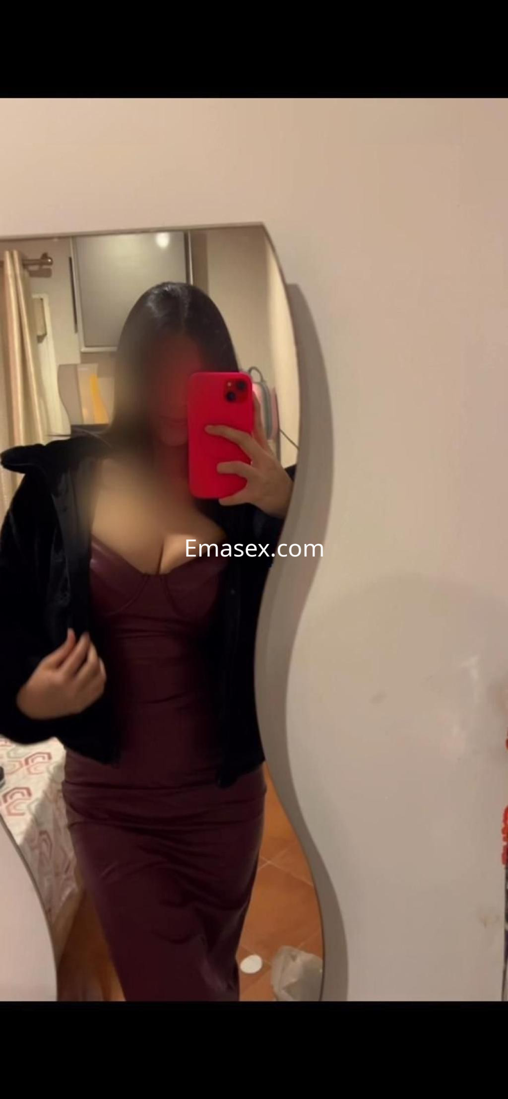 Escort en Huelva con vestido ajustado de color burdeos y abrigo oscuro, posando frente a un espejo.