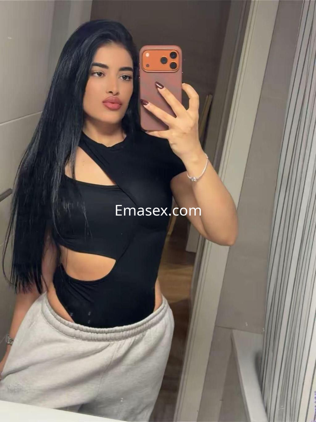 Puta en Madrid con cabello largo y negro, luciendo un body negro con cortes y pantalones grises en un espejo.