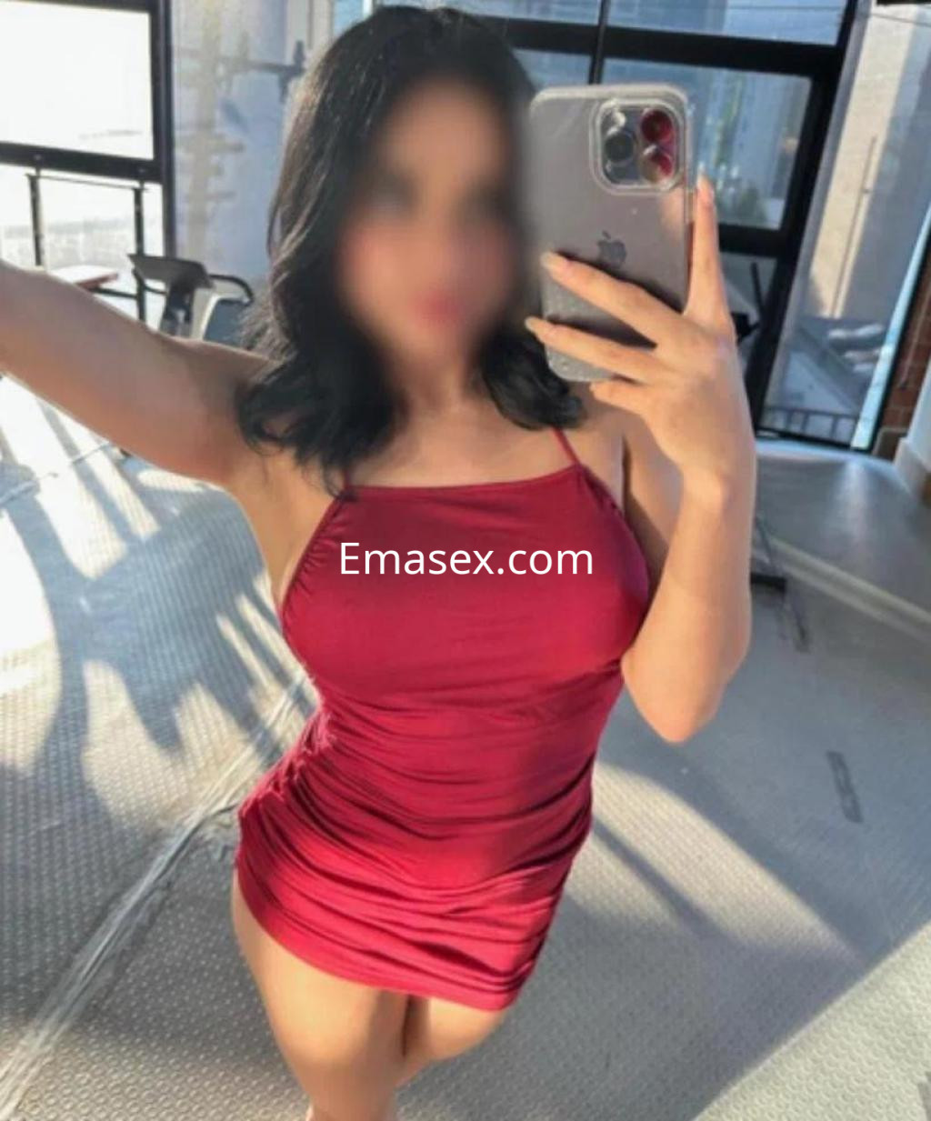 Escort en Pamplona posando con un vestido rojo ajustado, sosteniendo un teléfono móvil en un entorno luminoso.