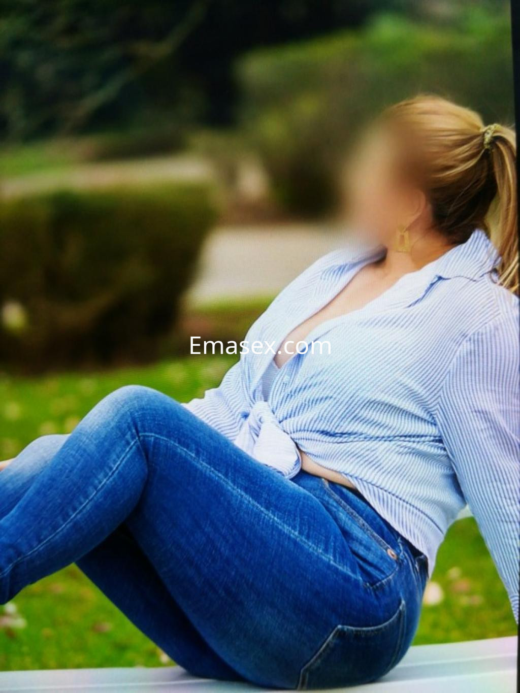 Escort en Vigo, con blusa de rayas y jeans ajustados, posando sentada en un entorno natural.