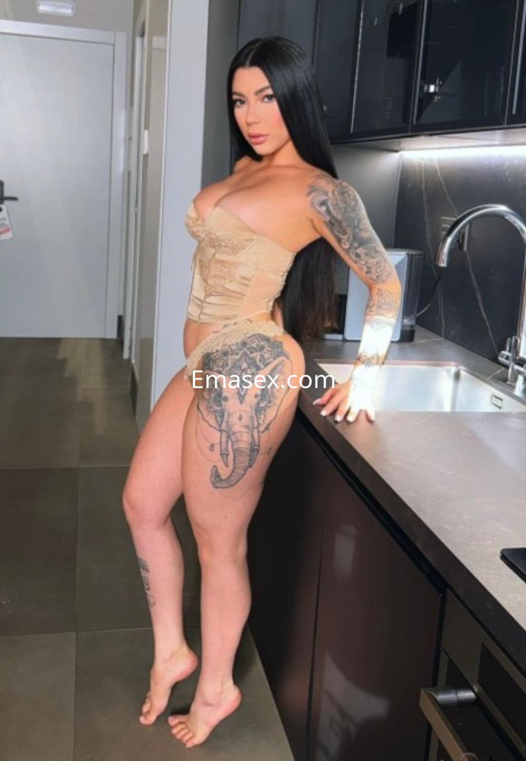 Puta de Almería posando en una cocina moderna, con un corset claro, tatuajes visibles y cabello lacio.