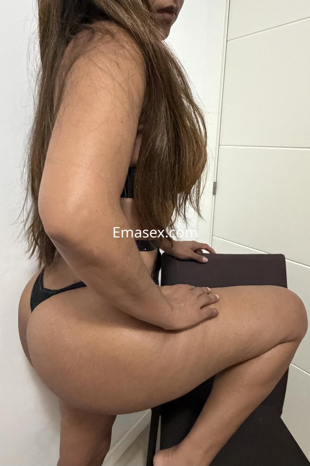 Escort en Tarragona posando de perfil, con lencería negra y cabello largo, sentada sobre una silla.