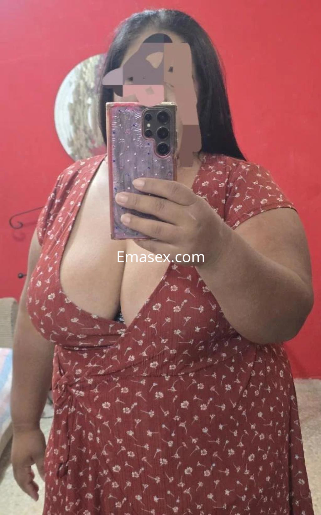 Escort en Marbella con vestido rojo con estampado floral, posando frente a un espejo en un ambiente acogedor.