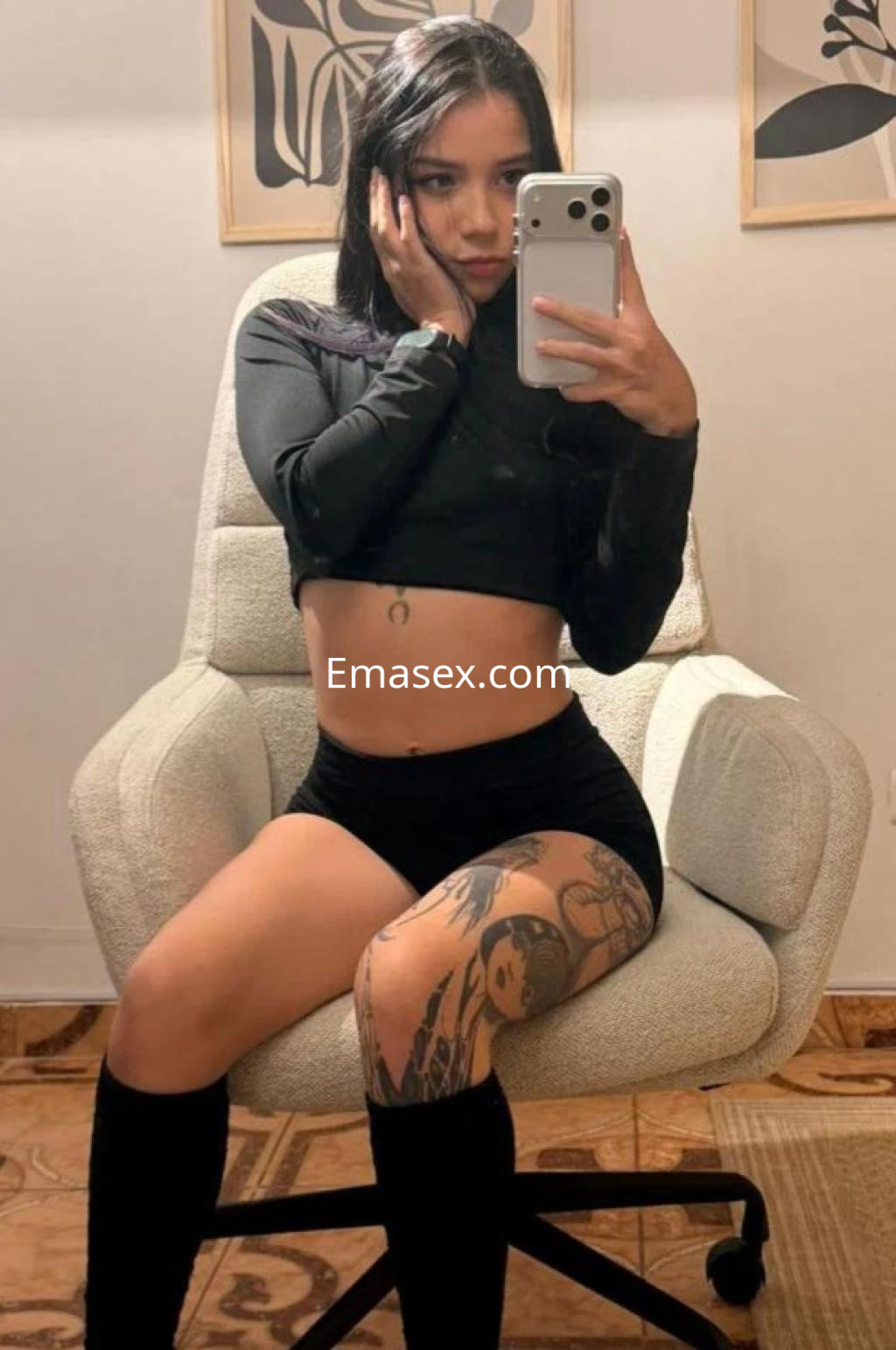 Escort en Málaga posando en un sillón, viste un top negro y shorts, mostrando un tatuaje en la pierna.