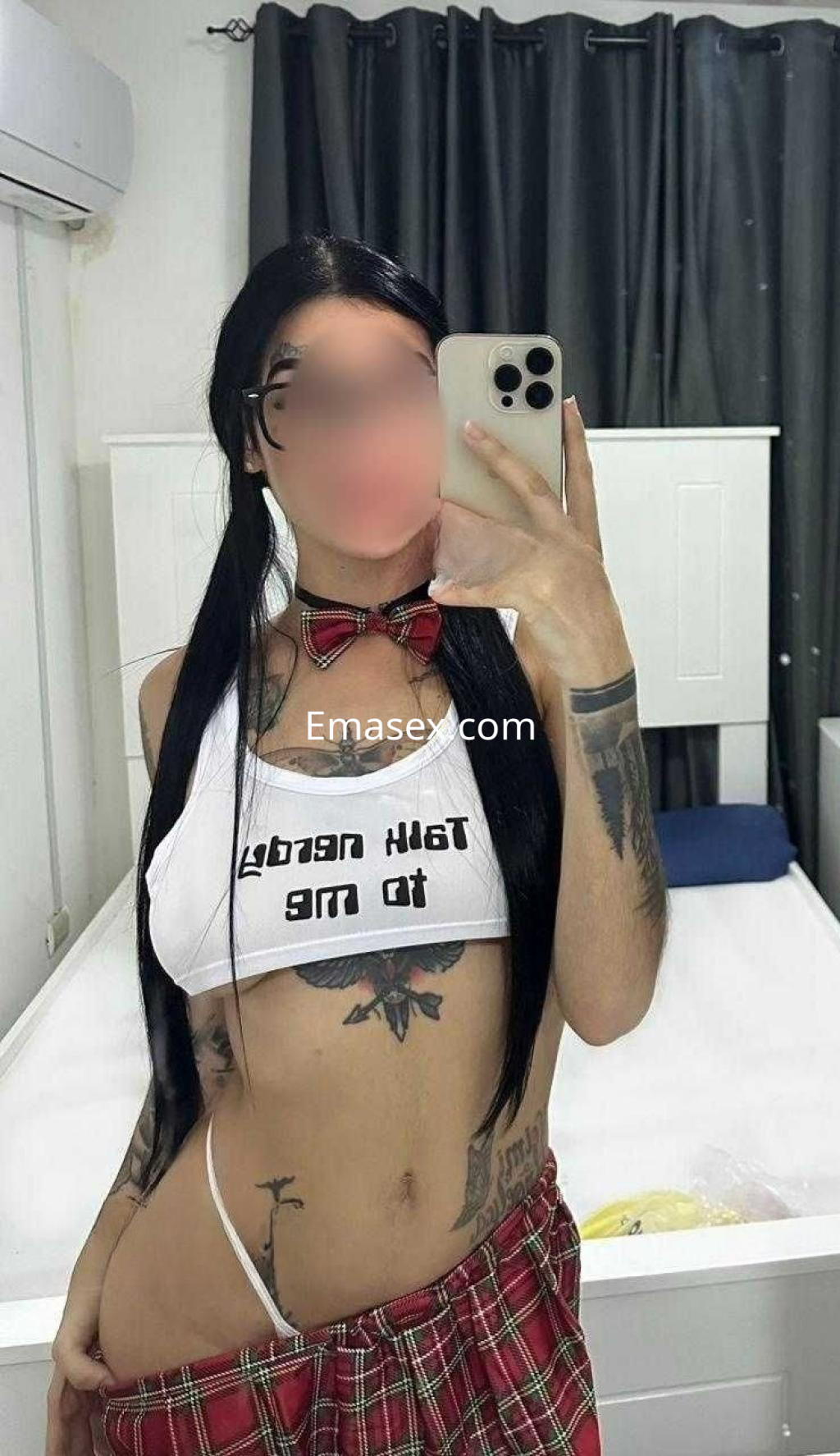 Escort en Zaragoza con cabello largo y lacio, viste un top blanco con texto y una falda a cuadros, mostrando tatuajes.