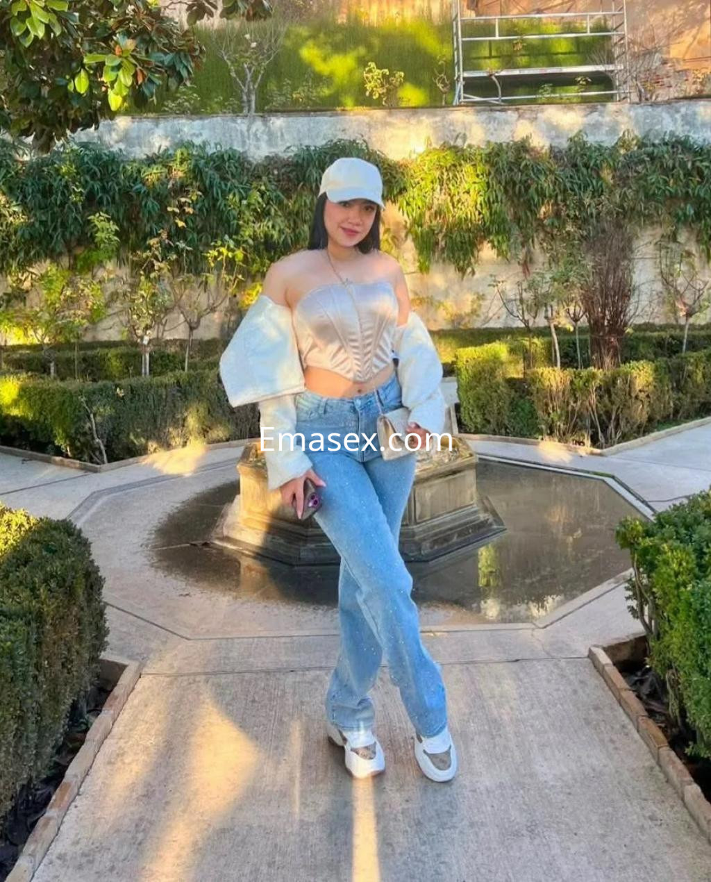 Escort en Zaragoza con top ajustado, chaqueta blanca y jeans claros, posando en un jardín con fuentes.