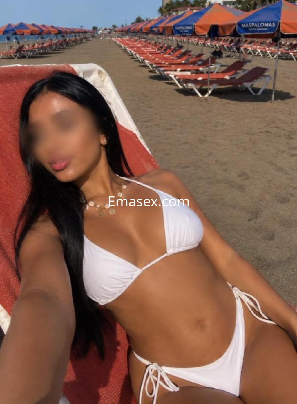 Escort en Las Palmas, sentada en una tumbona en la playa, vistiendo un bikini blanco y luciendo un collar.