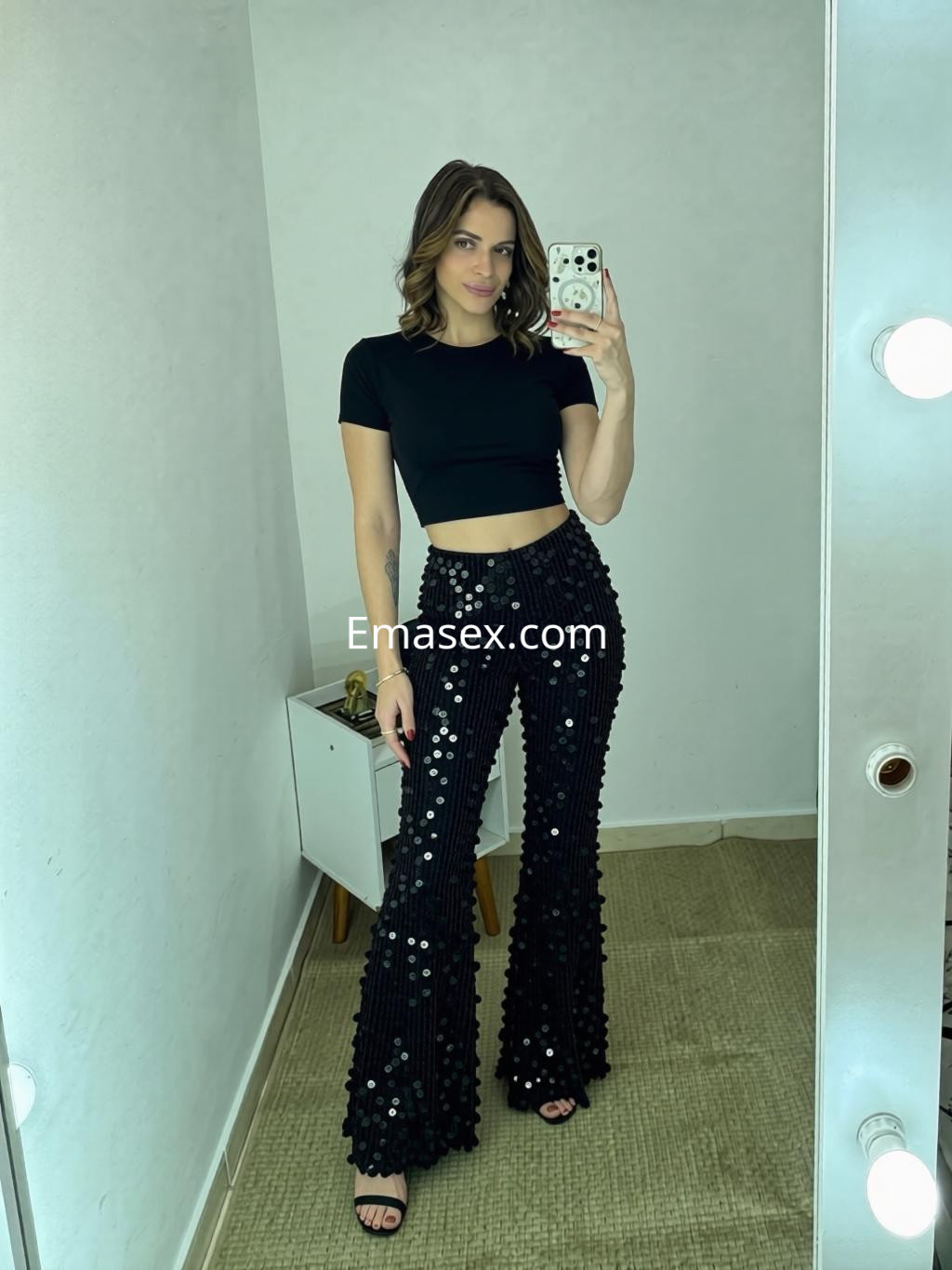 Escort en Santander posando en un espejo, vistiendo una blusa negra y pantalones brillantes de campana.