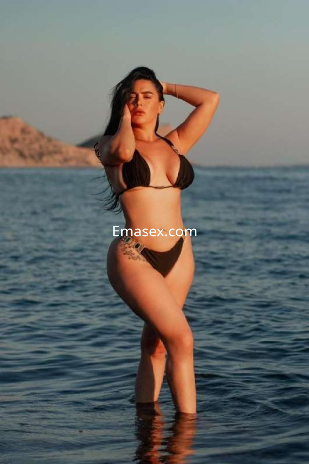 Escort en Ibiza vistiendo un bikini negro, posando en el agua con una mano en la cabeza y el sol de fondo.