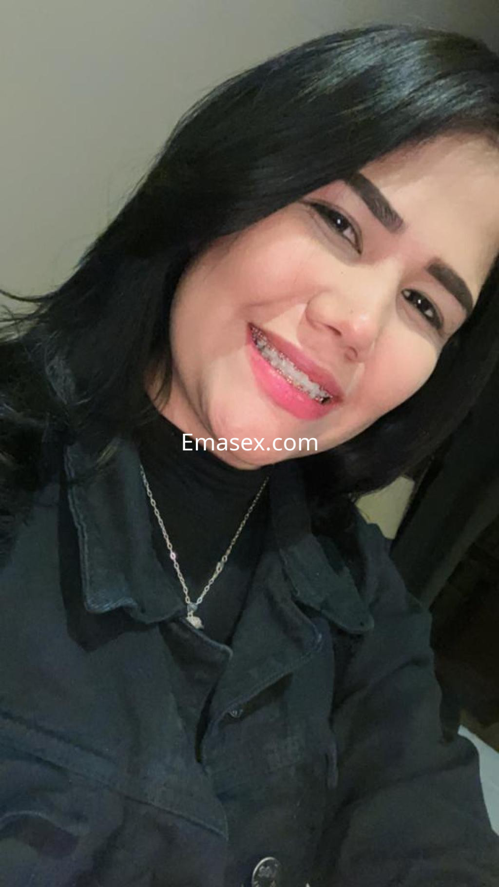 Escort en Coruña con cabello negro y lacio, sonriendo mientras viste una blusa negra y una cadena en el cuello.