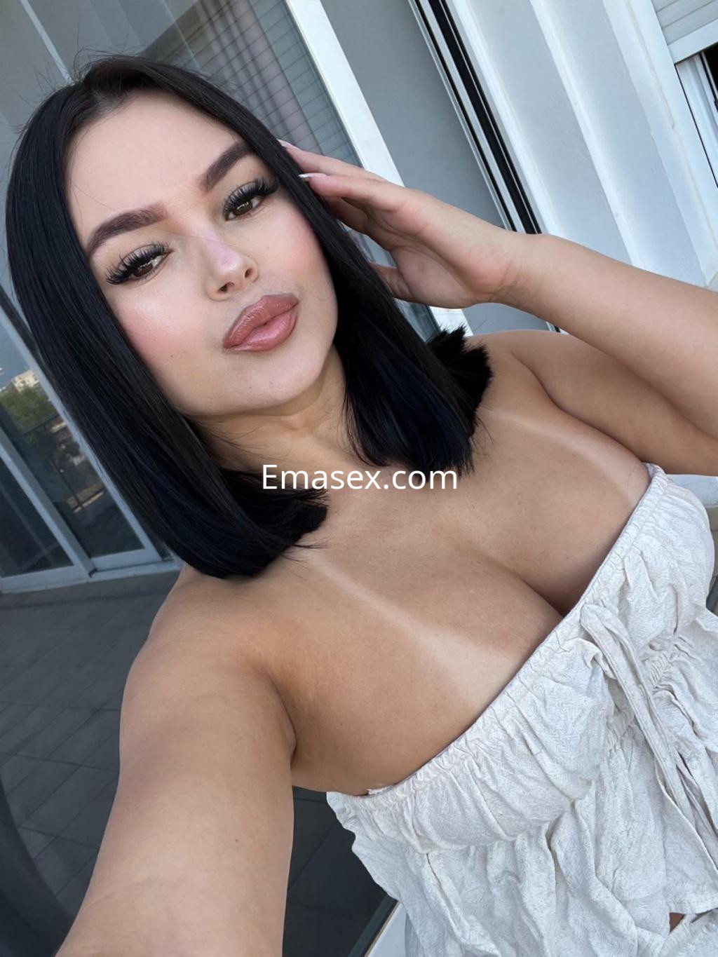 Escort en Barcelona con cabello negro lacio y un top claro, posando con una mano en la cara en un entorno luminoso.