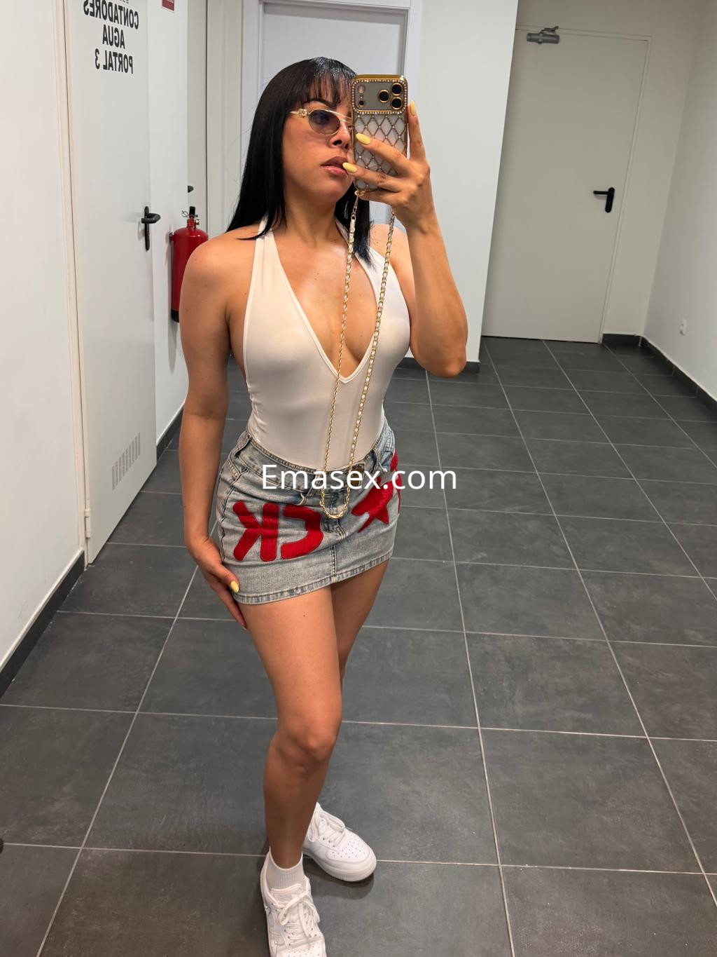 Escort en Vigo luciendo un body blanco y una falda de mezclilla con detalles rojos, posando frente al espejo.