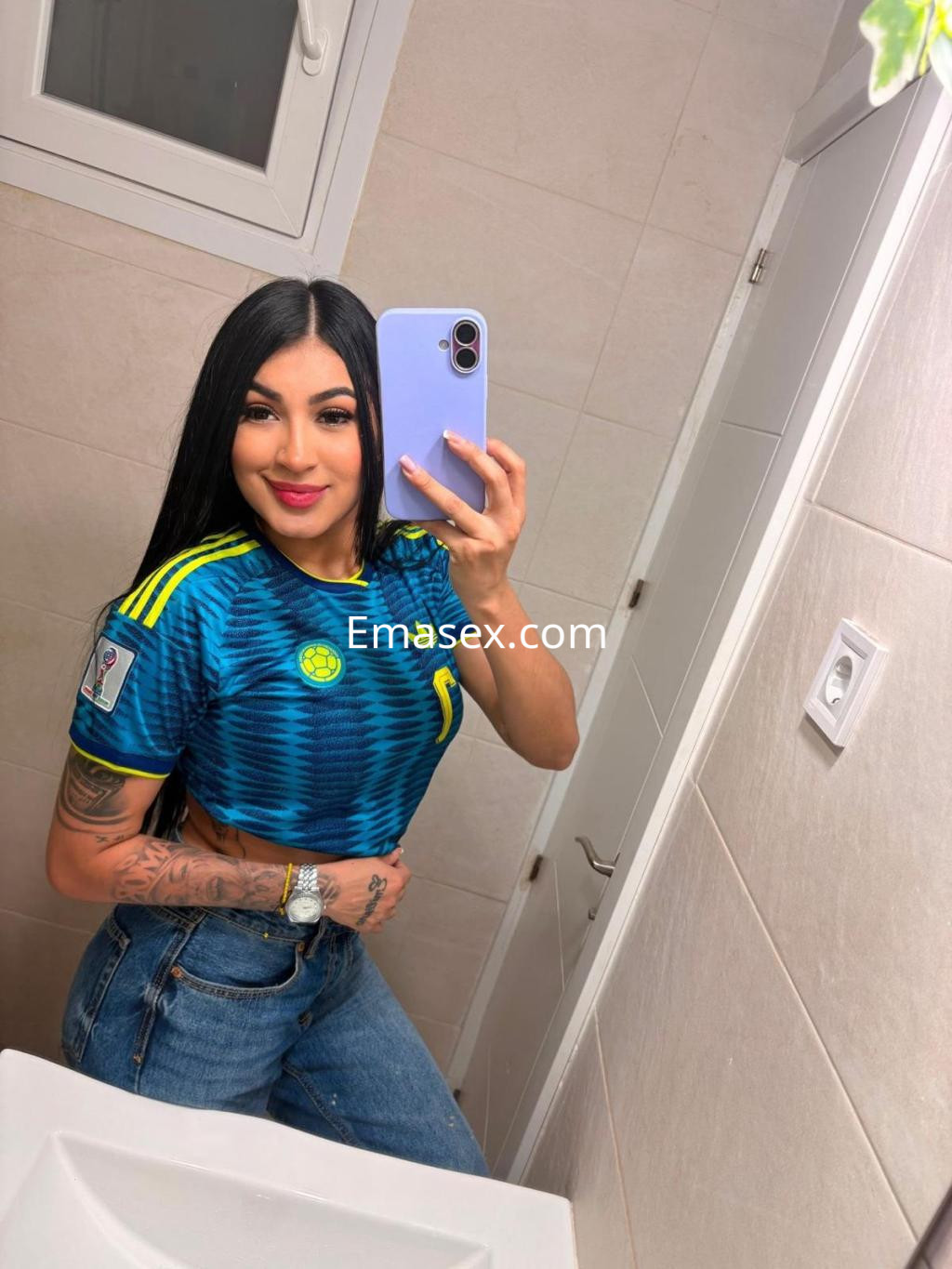 La escort en Bilbao posa en un baño, luciendo una camiseta deportiva azul y jeans ajustados mientras sonríe.