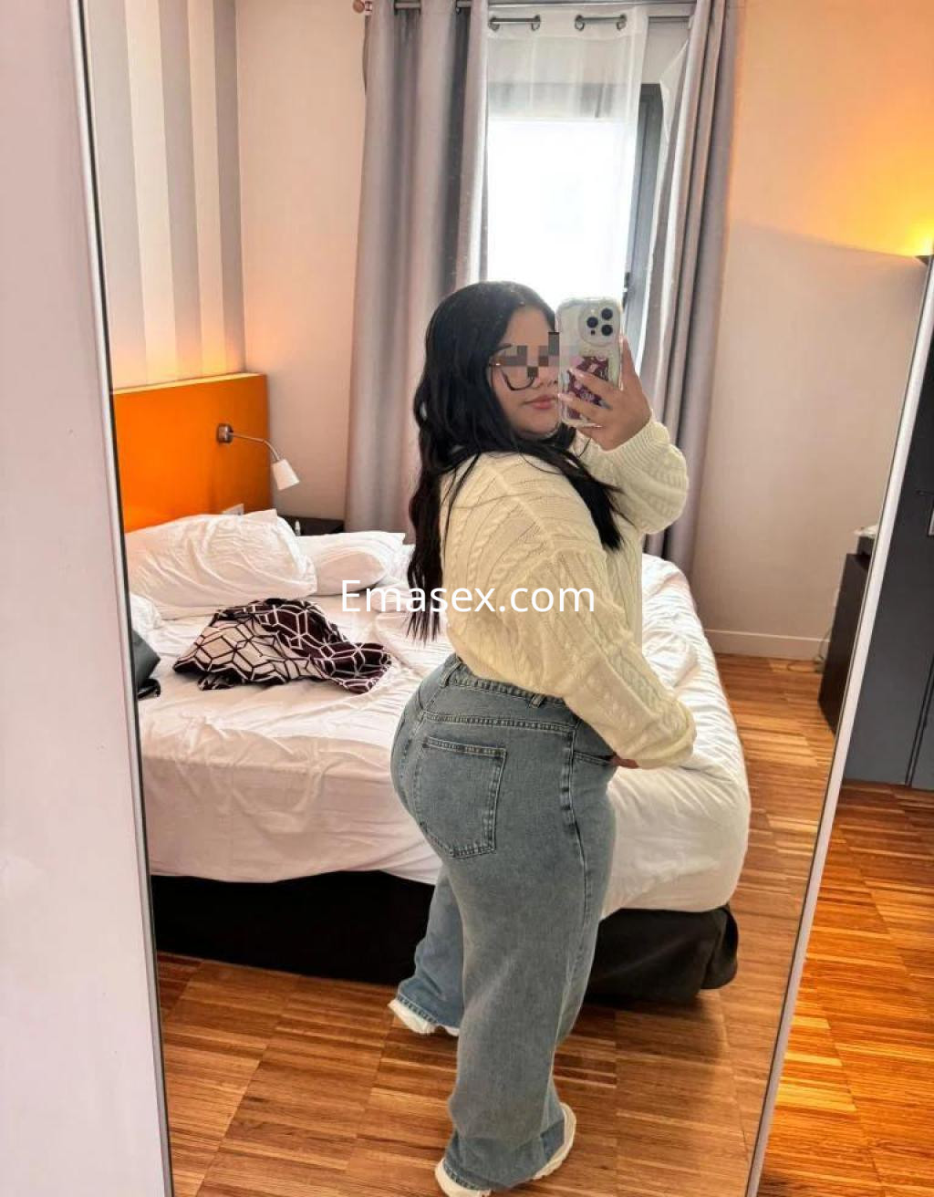 La puta posa en un espejo con un suéter blanco y jeans grises en una habitación de Alicante.