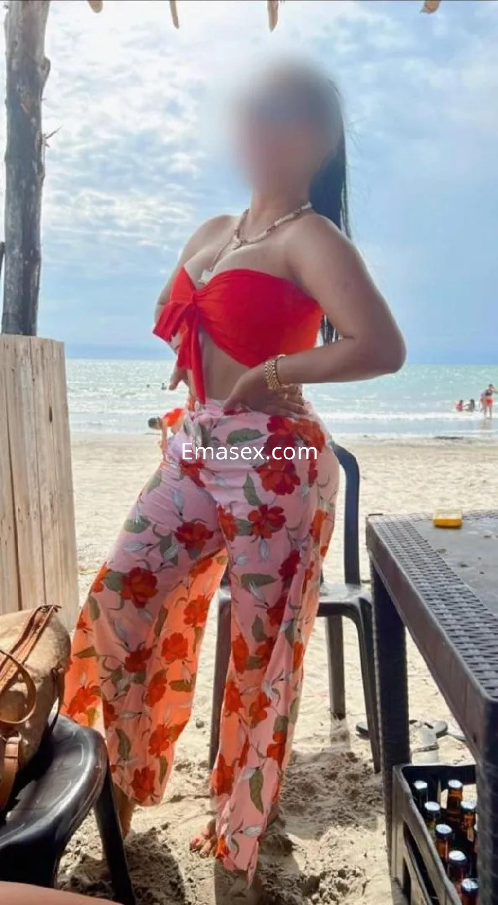 La puta en Barcelona posa en la playa, vistiendo un top rojo y pantalones florales, con el mar de fondo.