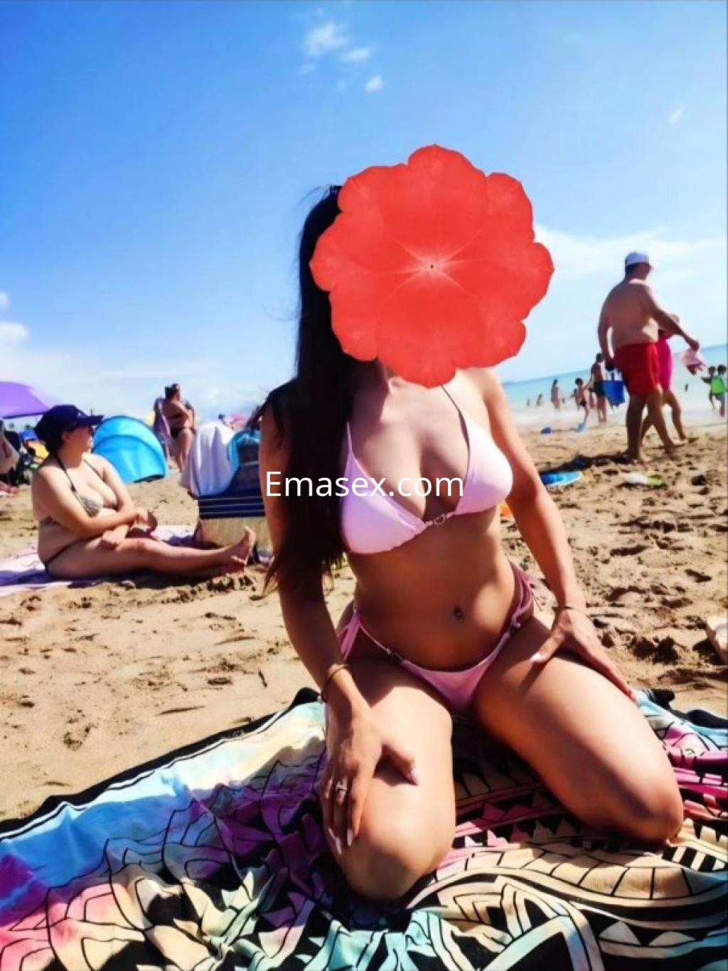 Puta en la playa de Coruña, usando un bikini rosa, de rodillas sobre una toalla en la arena, con un fondo soleado.