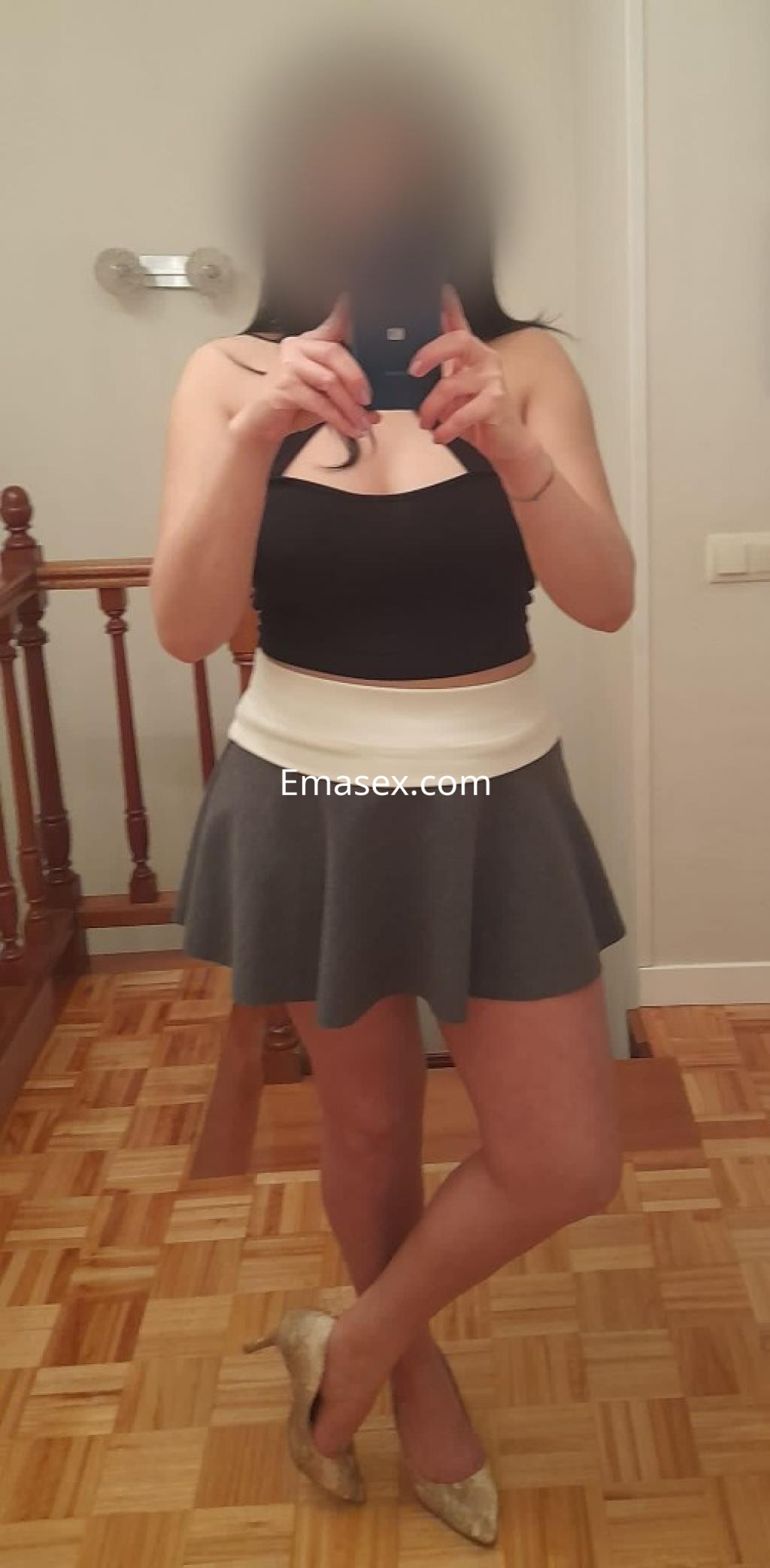 Escort en Oviedo con blusa negra sin mangas y falda corta gris, posando frente a un espejo.
