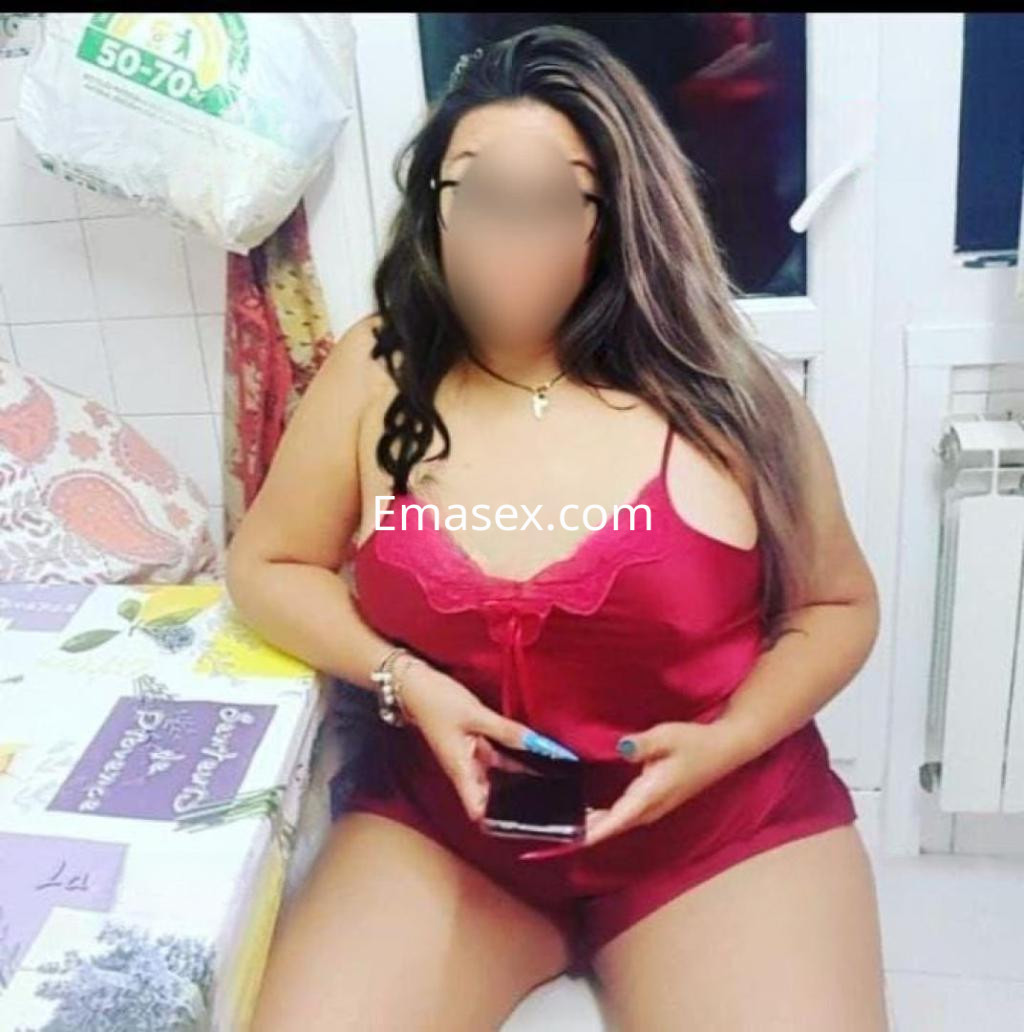 Puta en Oviedo luce un conjunto de lencería roja, sentada en una mesa con una expresión relajada.