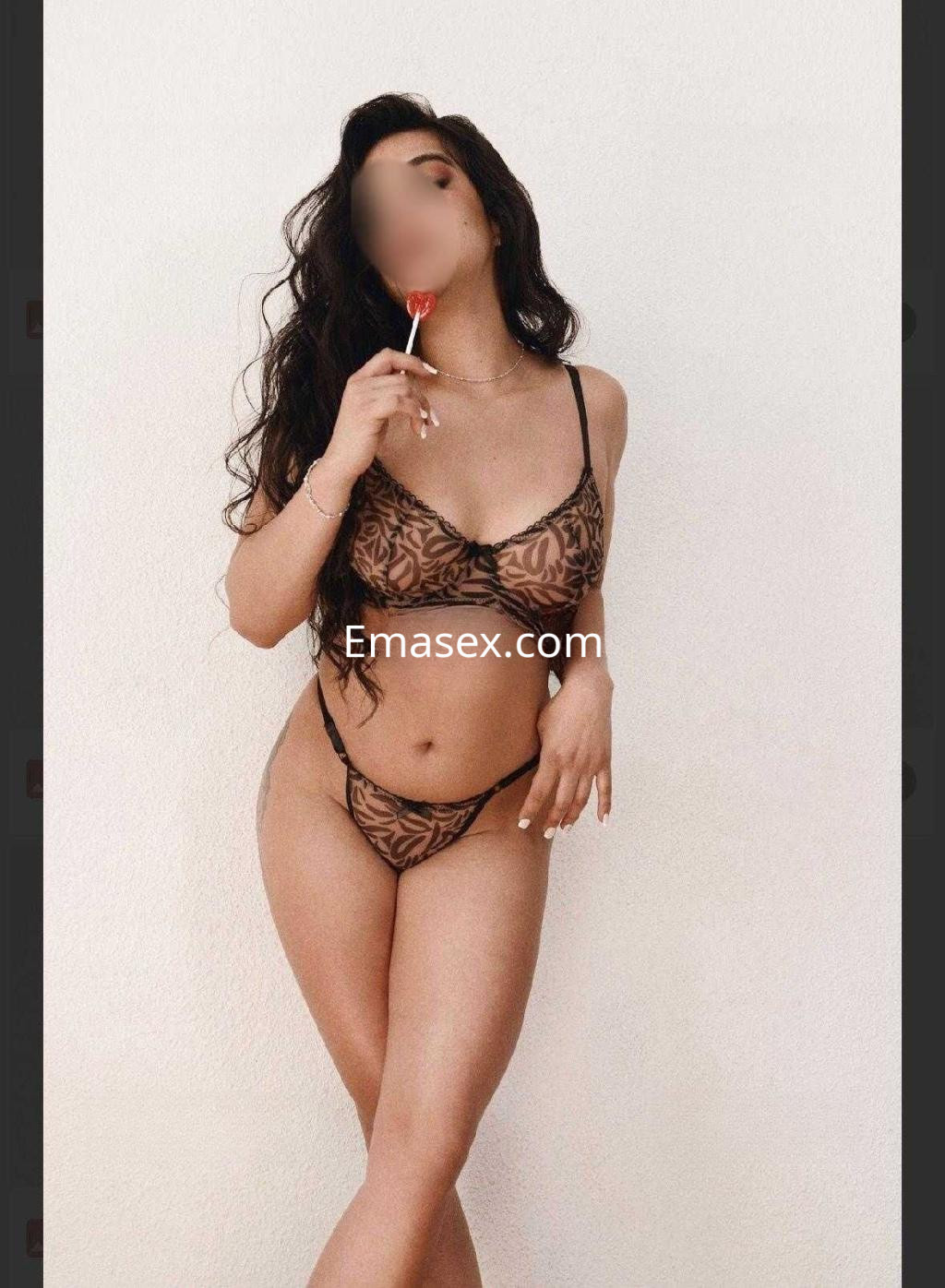 Escort en Toledo posando en lencería negra con estampado, sosteniendo un caramelo, con cabello largo y rizado.