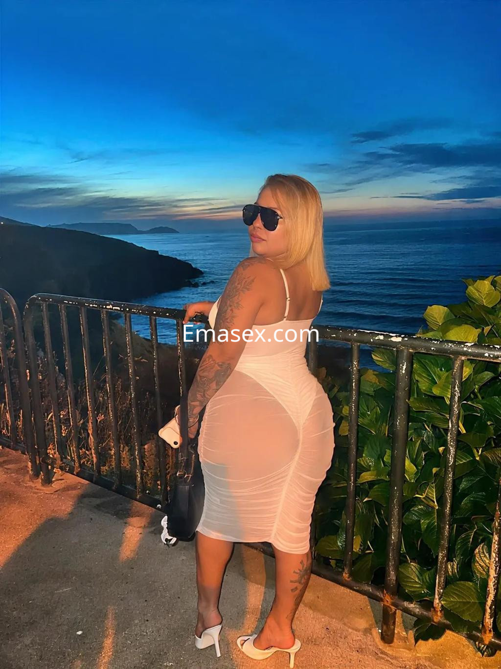 La puta lleva un vestido blanco ajustado y sandalias, posando al atardecer junto al mar en un paisaje costero.