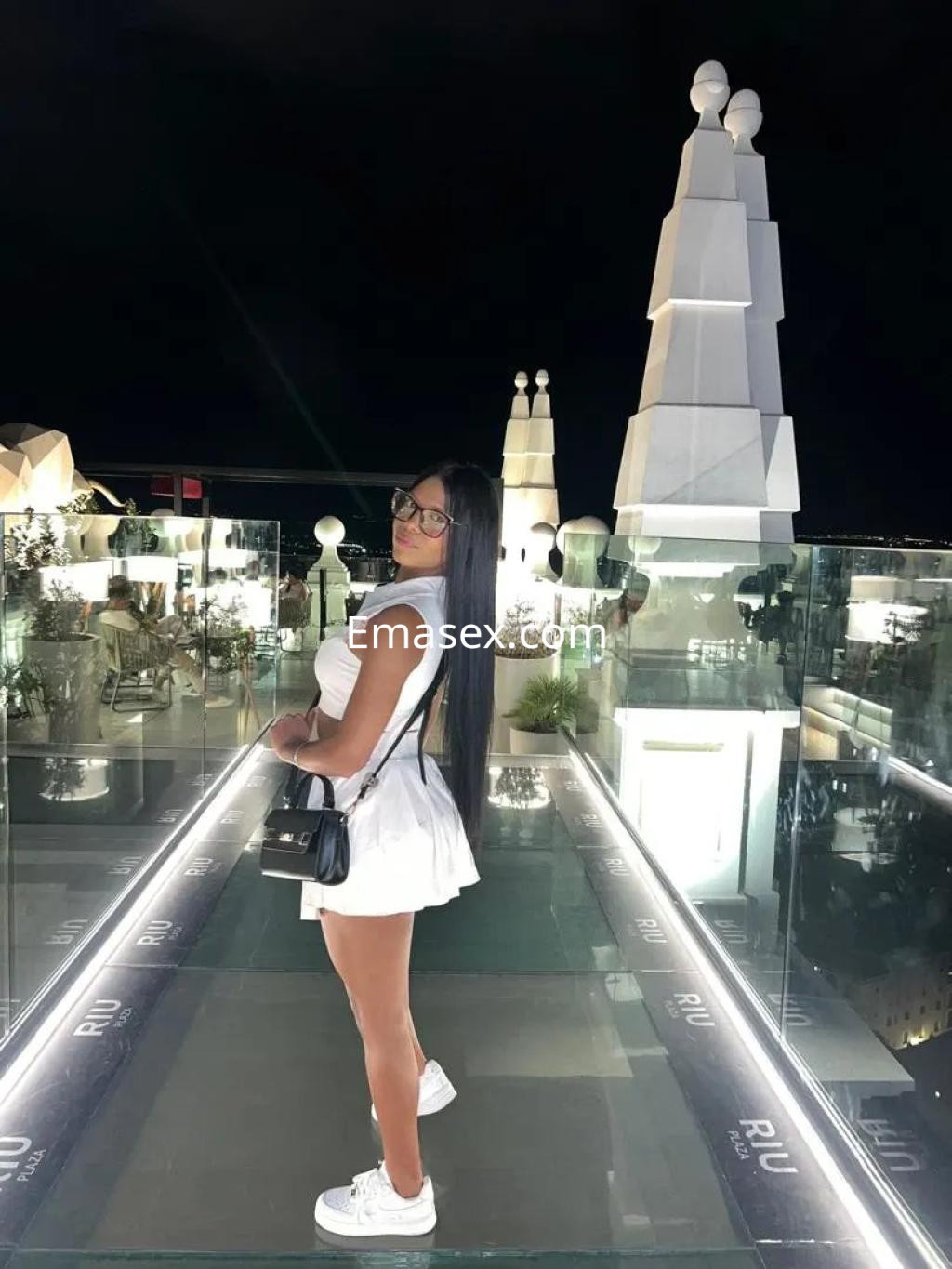 Escort en Valladolid, posando con un vestido blanco y gafas, en un entorno moderno y elegante al aire libre.