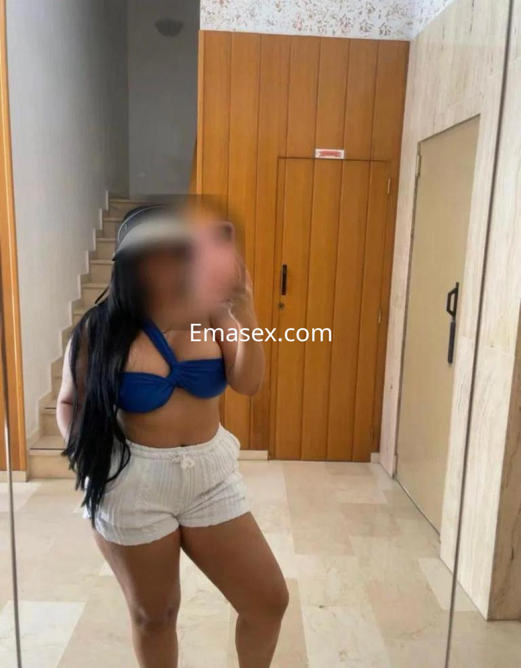 Escort en Valladolid, posando frente a un espejo, con un top azul y pantalones cortos blancos.