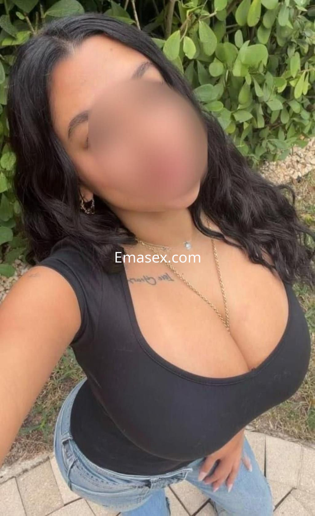La escort en Zaragoza lleva una camiseta negra ajustada y jeans, posando con un fondo de vegetación.