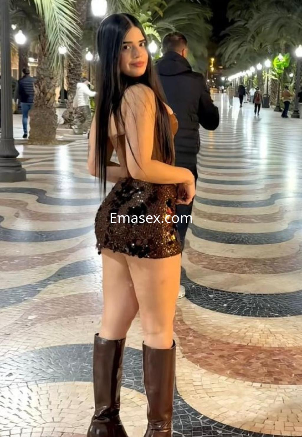 Puta de Almería posa de espaldas en una calle pavimentada, luciendo un vestido corto brillante y botas altas.