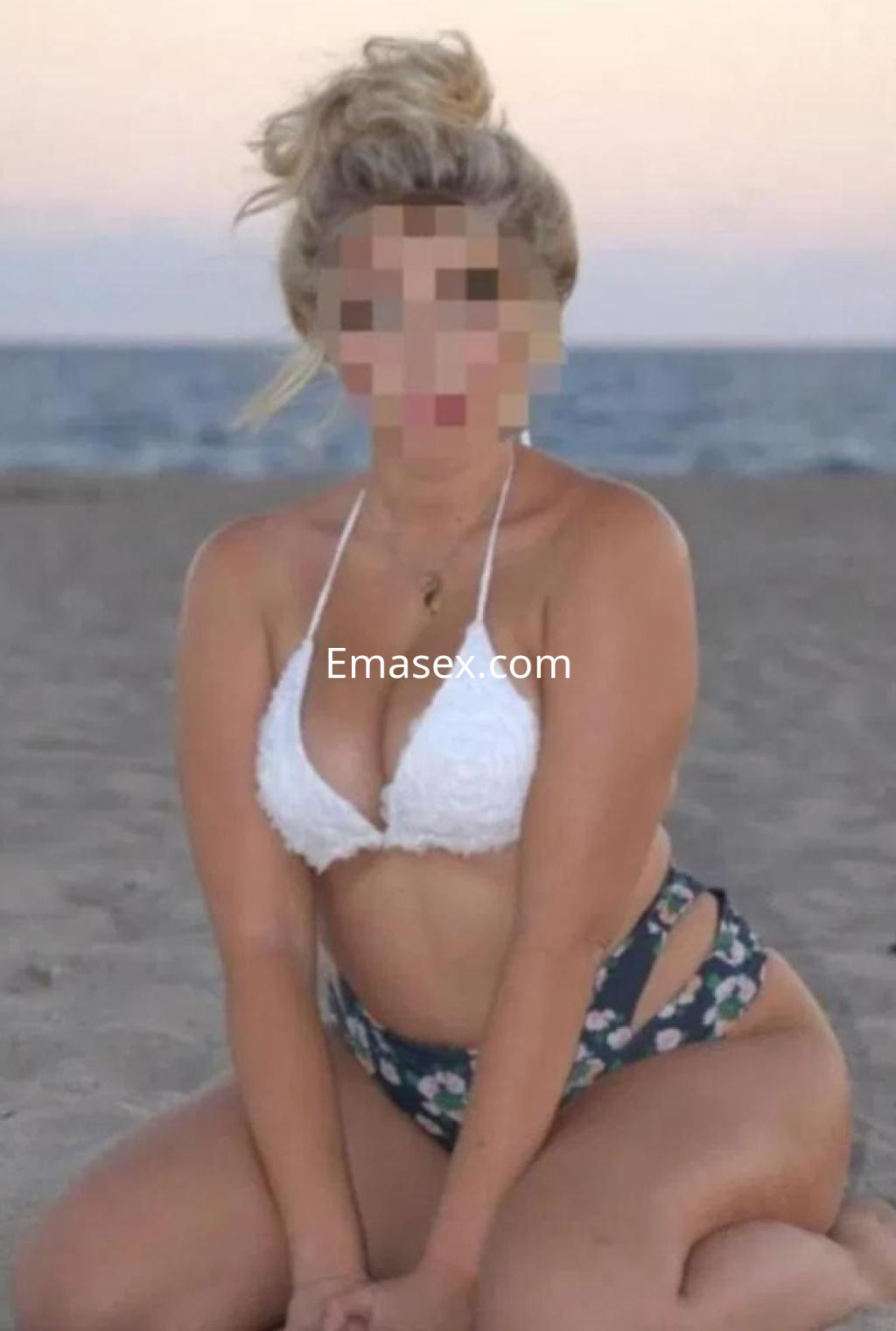 Escort en Málaga, vestida con un bikini blanco y pantalones de flores, posando en la playa al atardecer.