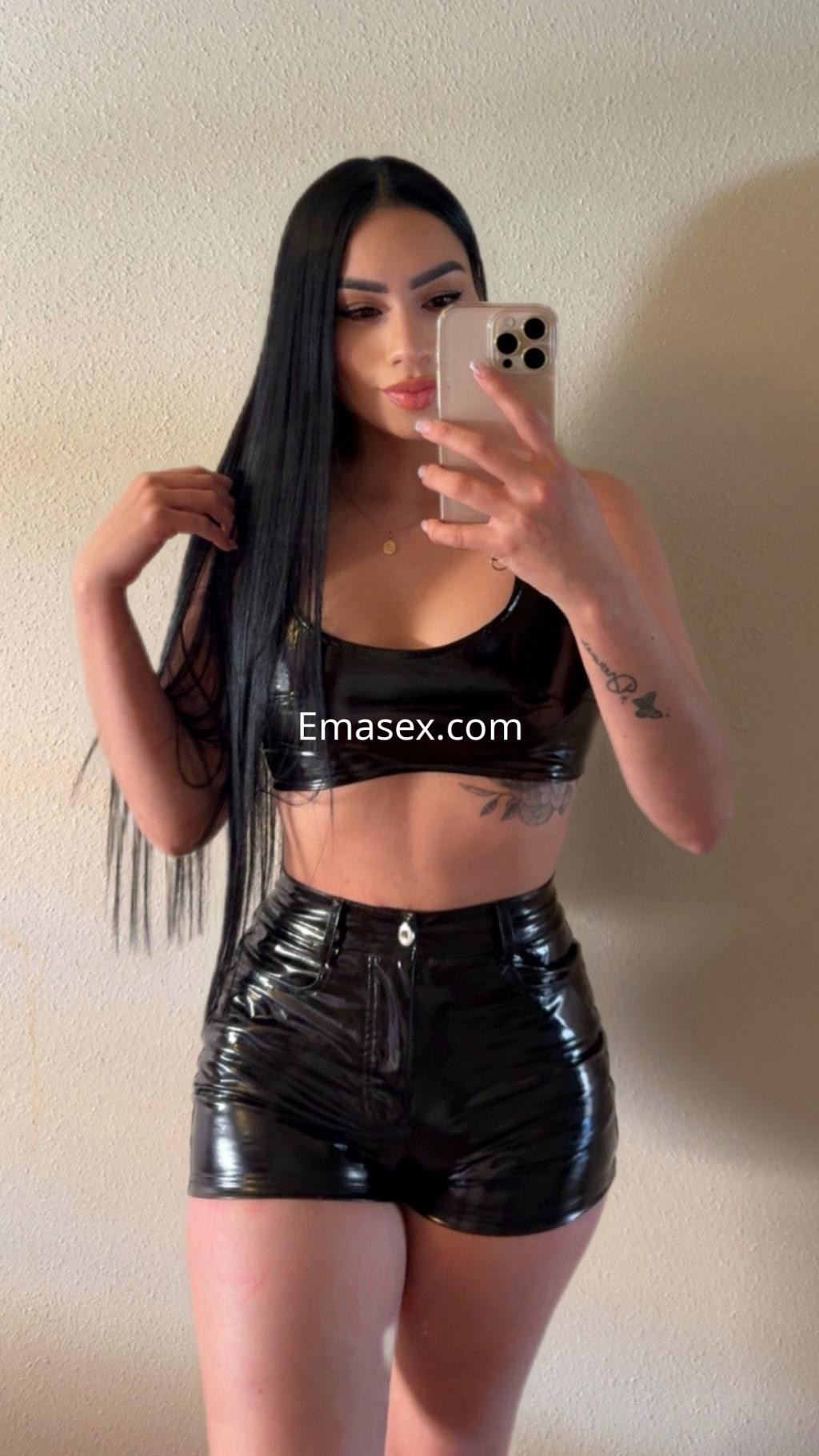 Escort en Donosti con cabello lacio y negro, vistiendo un crop top y shorts de vinilo negro, posando frente a un espejo.