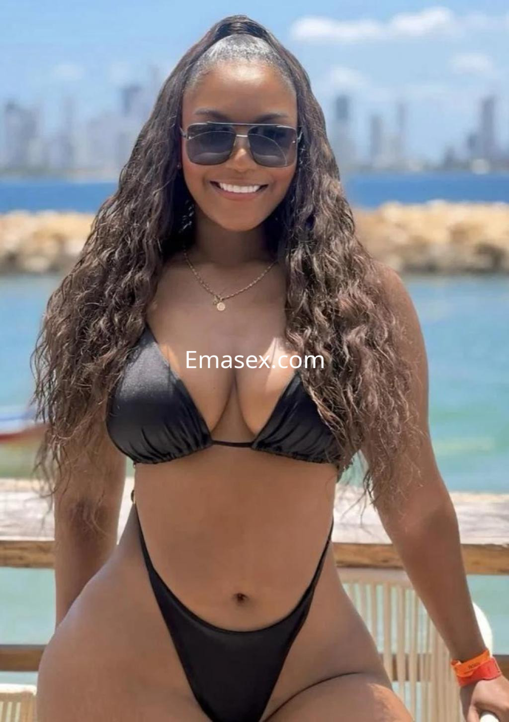 Escort en Granada con cabello rizado y oscuro, luciendo un bikini negro y gafas de sol frente al mar.
