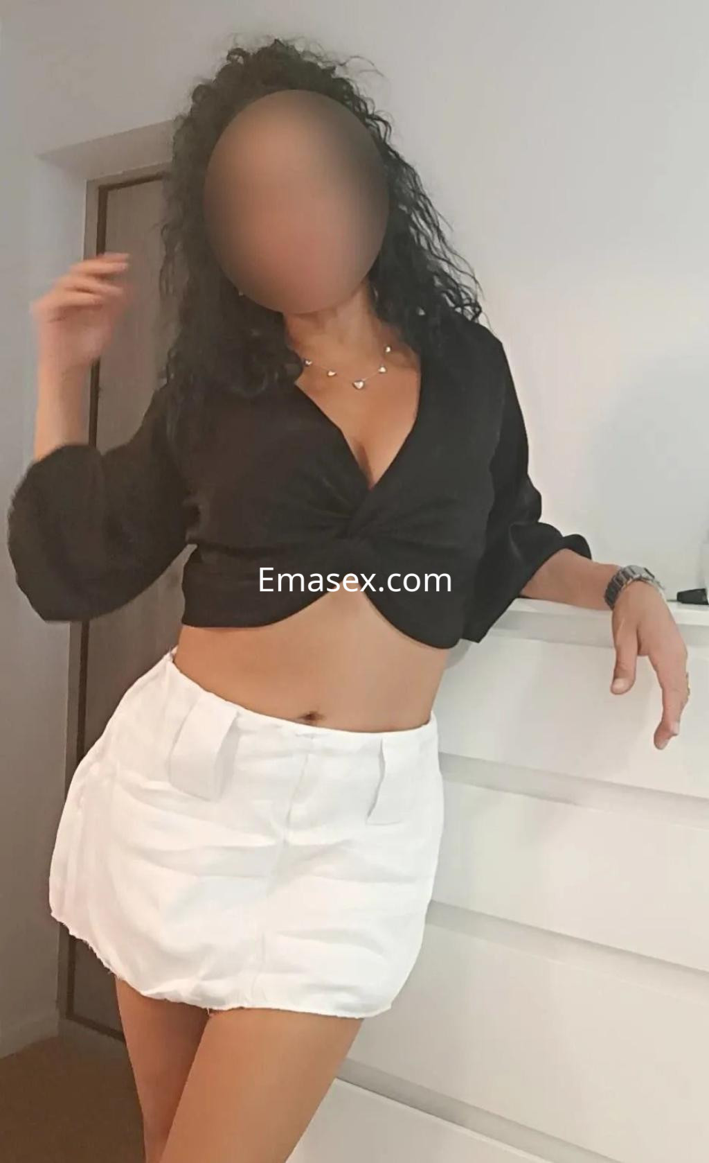 Escort en Sevilla luciendo un top negro ajustado y una falda corta blanca, con cabello rizado y suelto.