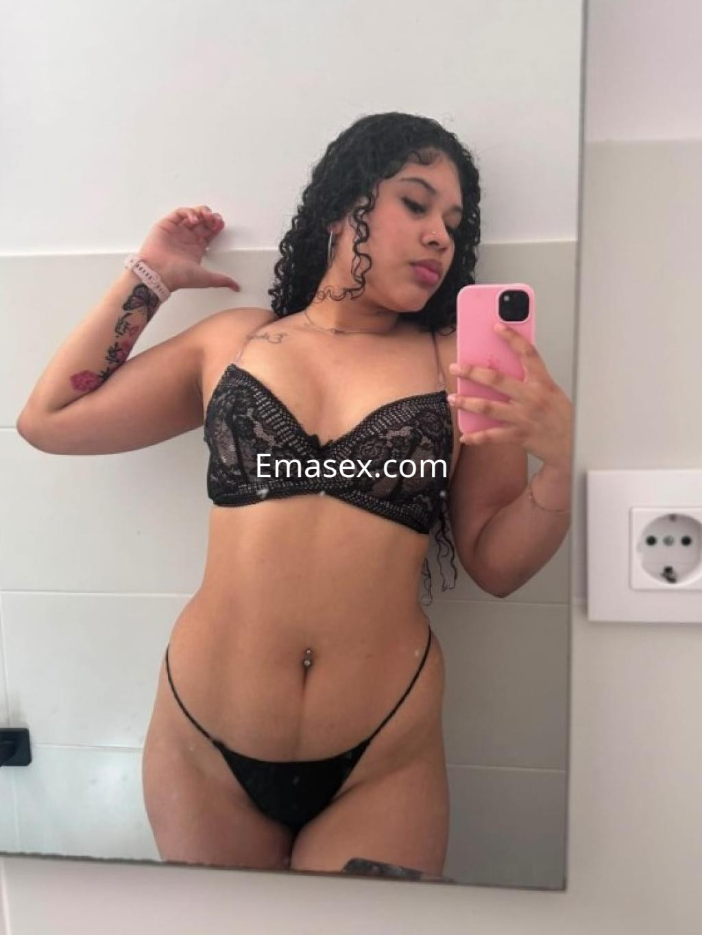 Escort en Lleida posando en un espejo, lleva un conjunto de lencería negra y muestra su tatuaje en el brazo.
