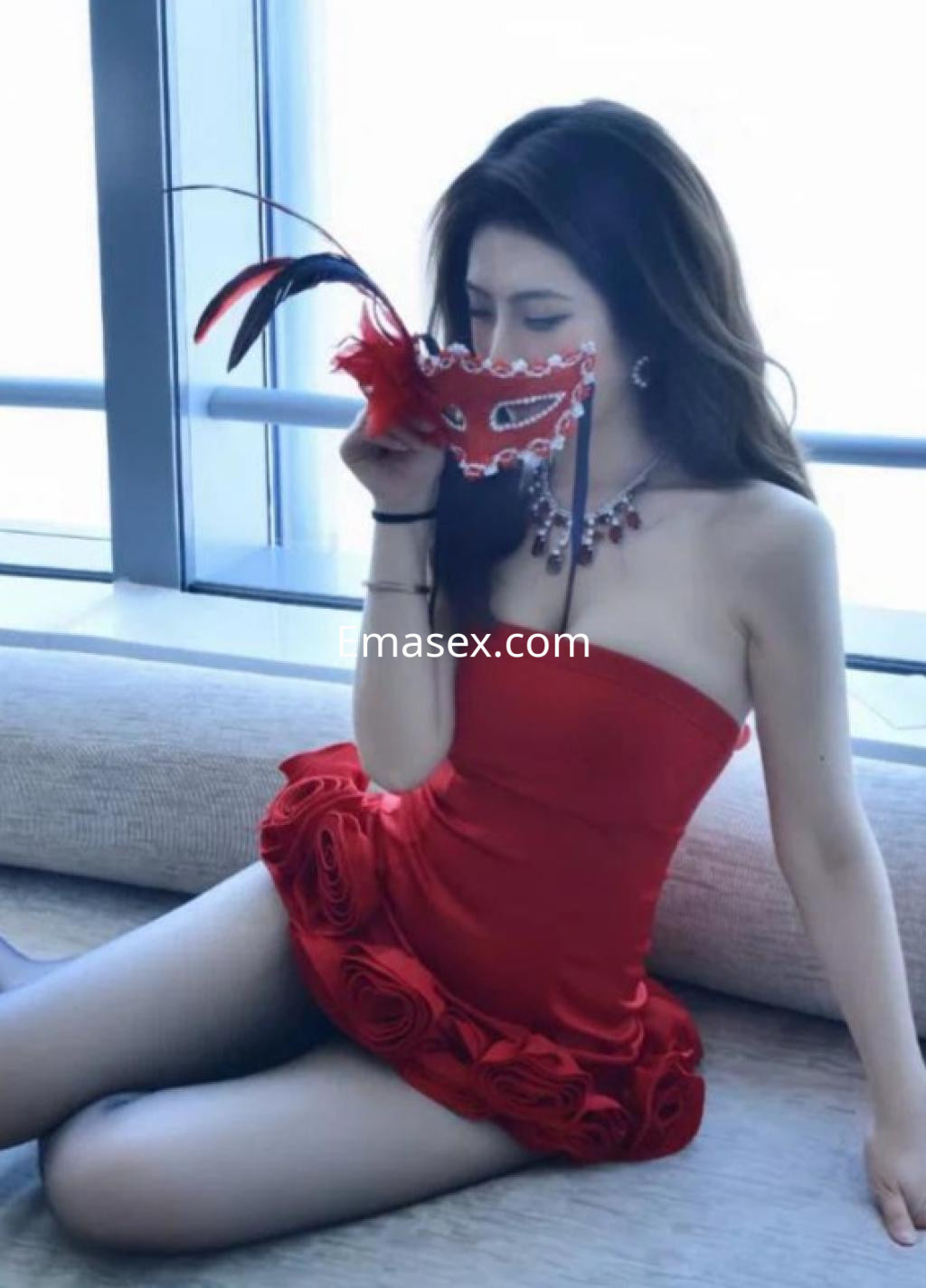 Escort en Girona con vestido rojo ceñido, adornos en la parte inferior y máscara decorativa, posando sentada.