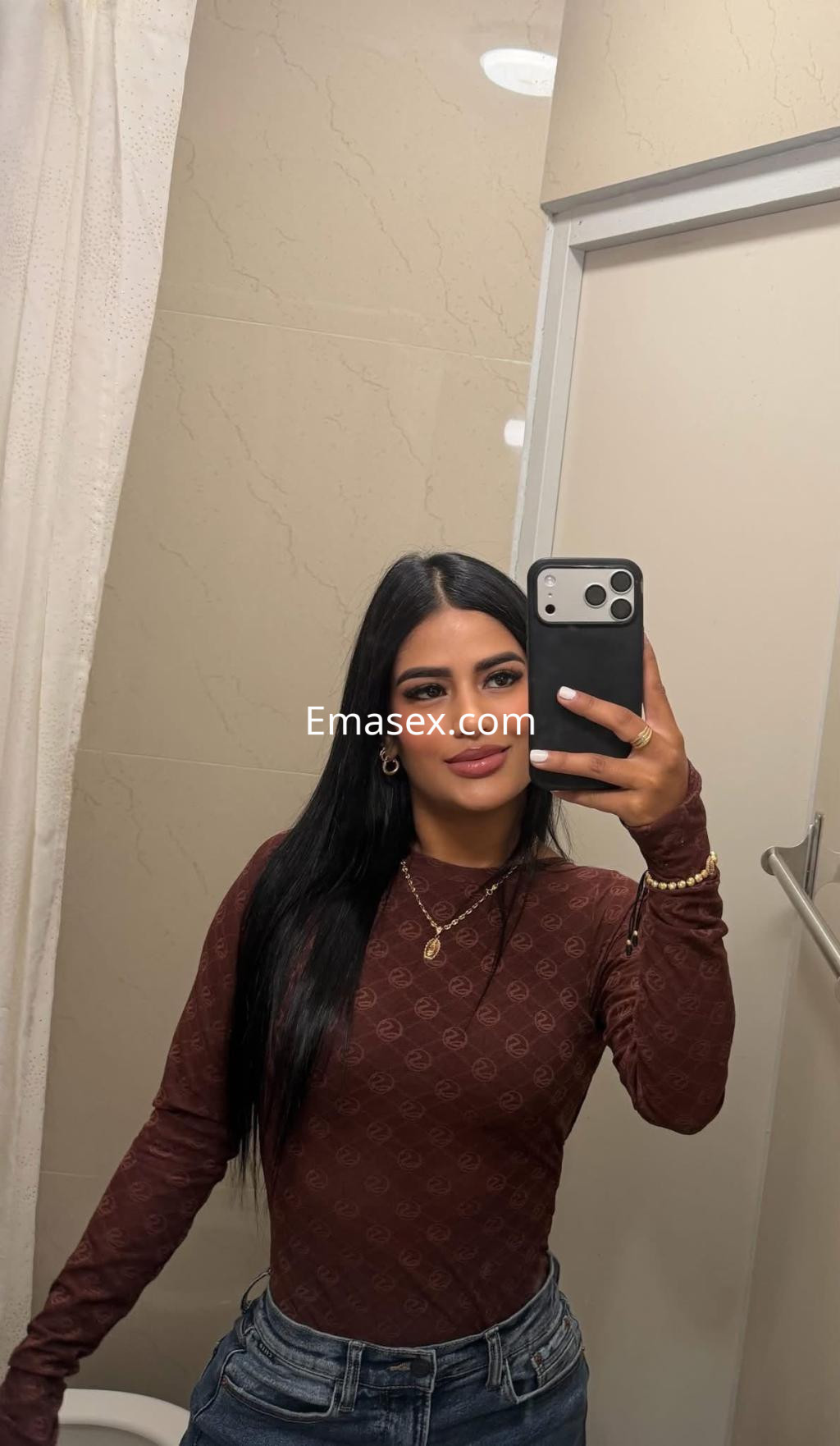 Puta de Bilbao posando en un baño con una blusa marrón ajustada y jeans, mostrando una sonrisa confiada.