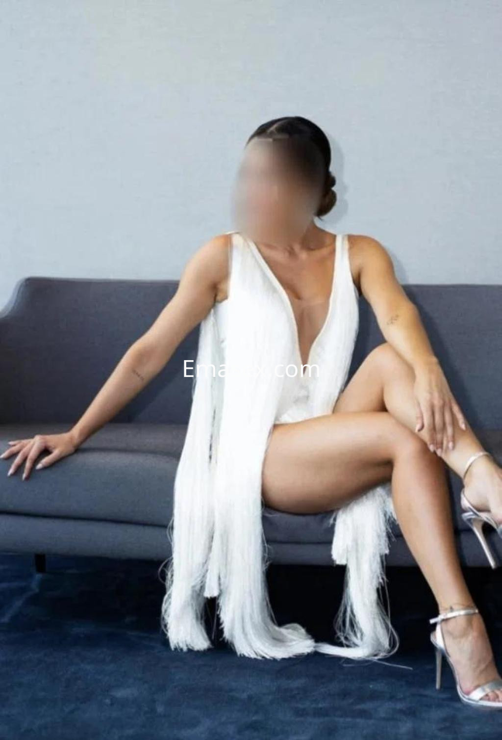 Puta de Almería sentada en un sofá, vistiendo un conjunto blanco con flecos y tacones plateados.