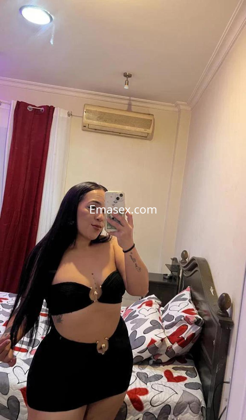 Escort en Badajoz posando en un espejo, lleva un conjunto negro y se encuentra en un dormitorio decorado con corazones.