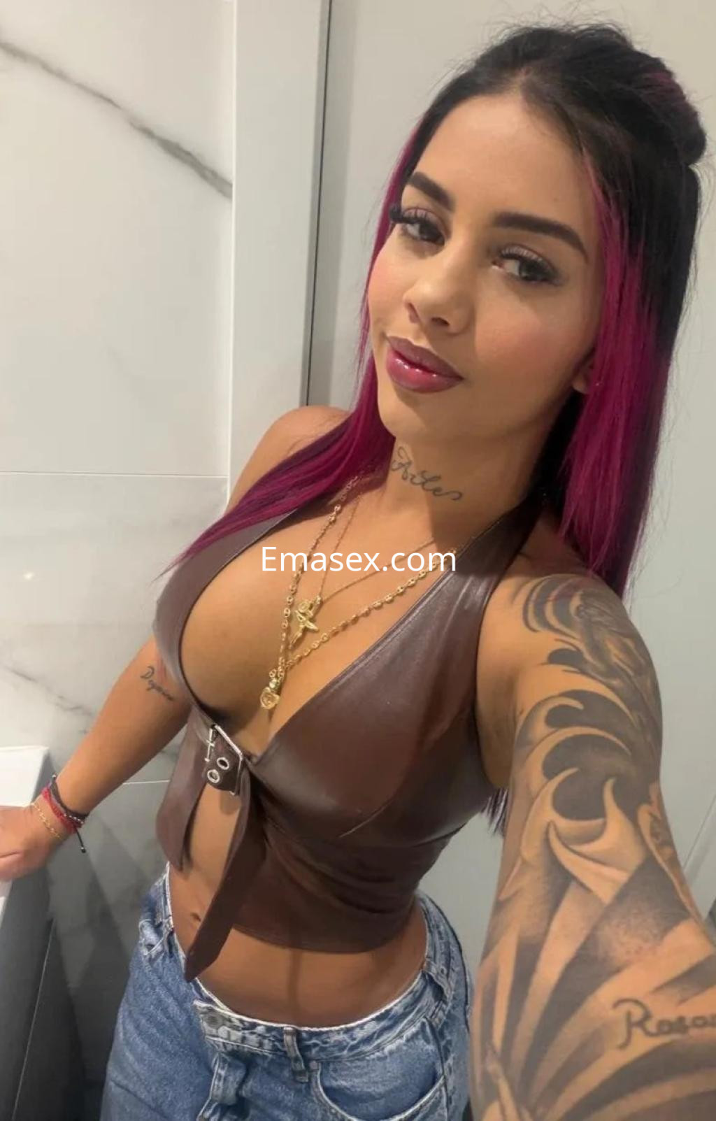 La puta luce un top de cuero marrón con escote y jeans, mostrando tatuajes y cabello rosa en Málaga.