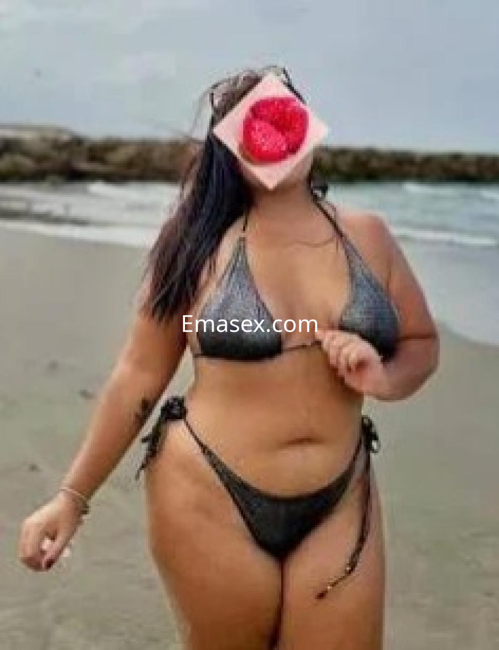 Escort en Granada con bikini negro en la playa, sosteniendo una rodaja de sandía frente a su rostro.