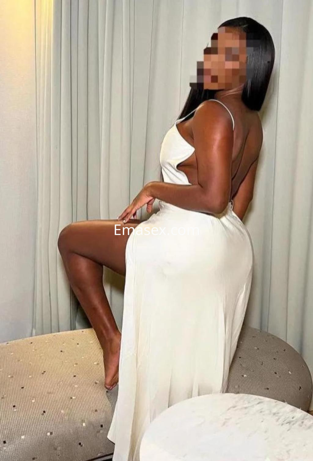 Escort posando de perfil en Sevilla, con un vestido blanco de tirantes y una pierna al descubierto, sentada en un sofá.