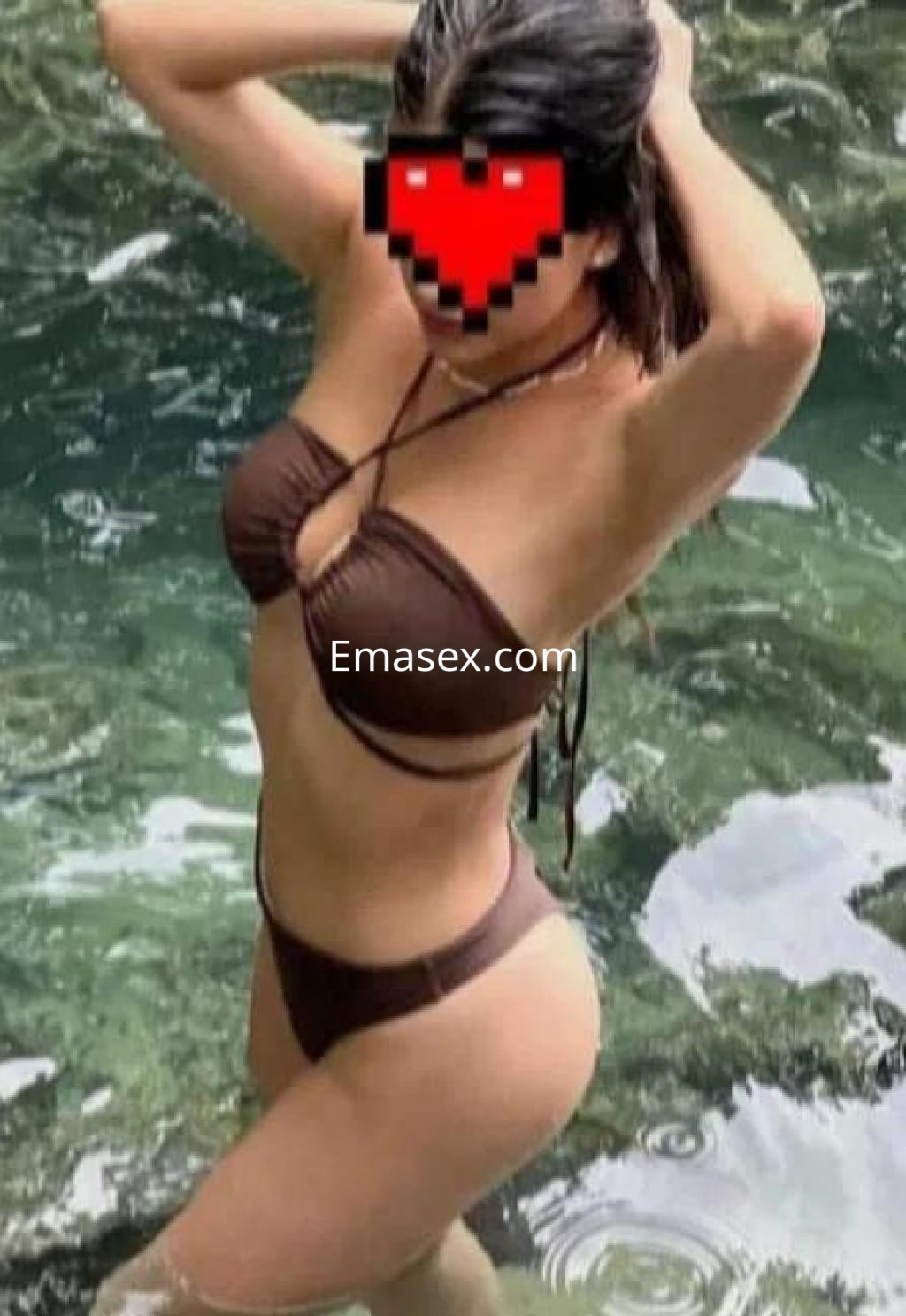 Escort en Lleida, posando en el agua con un bikini marrón, sujetando su cabello con una mano.